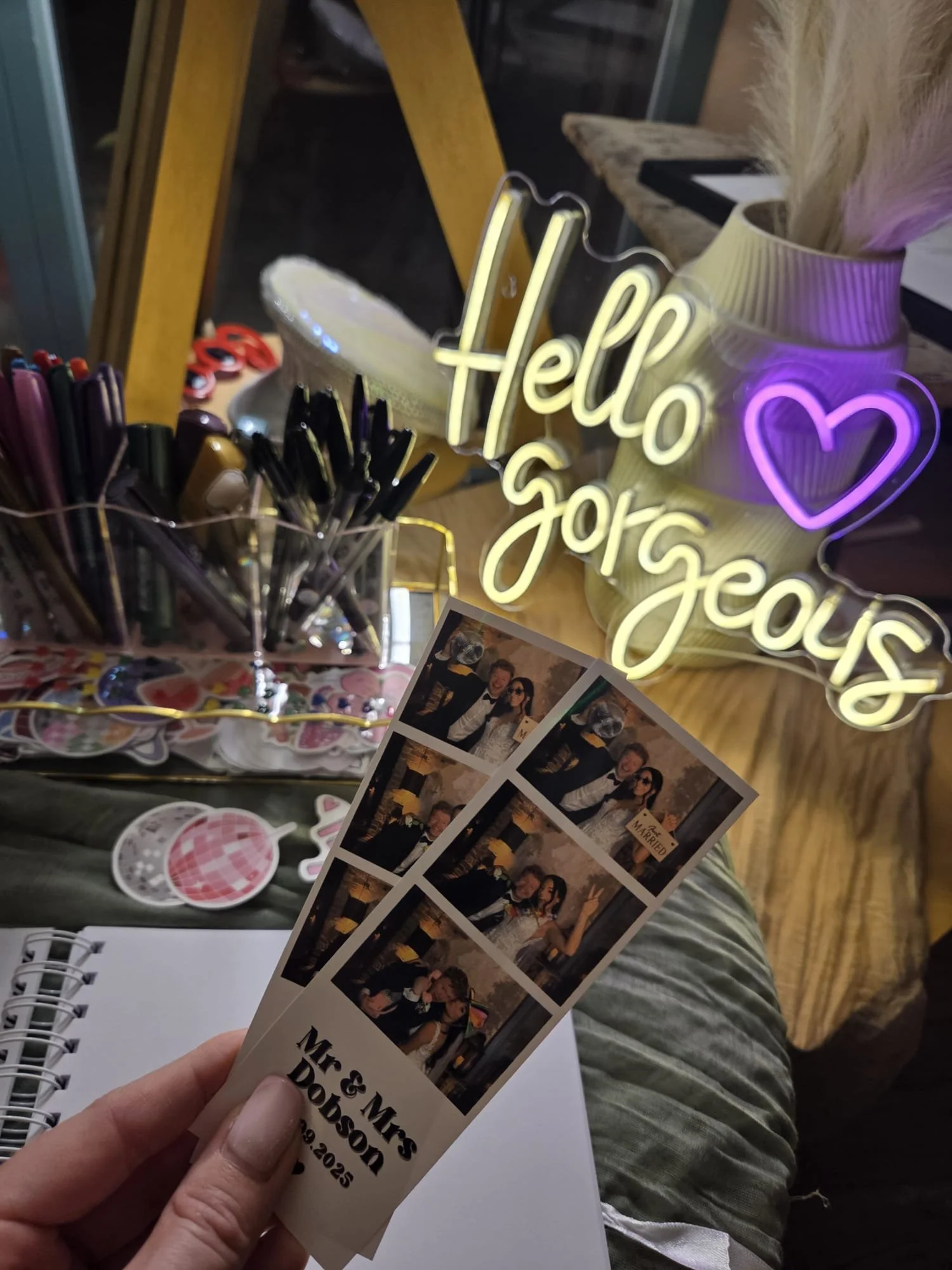 Photo of photo booth strips with wedding pics, a neon sign that says 'Hello George,' a vase, and a container with markers and pens, on a wooden table.