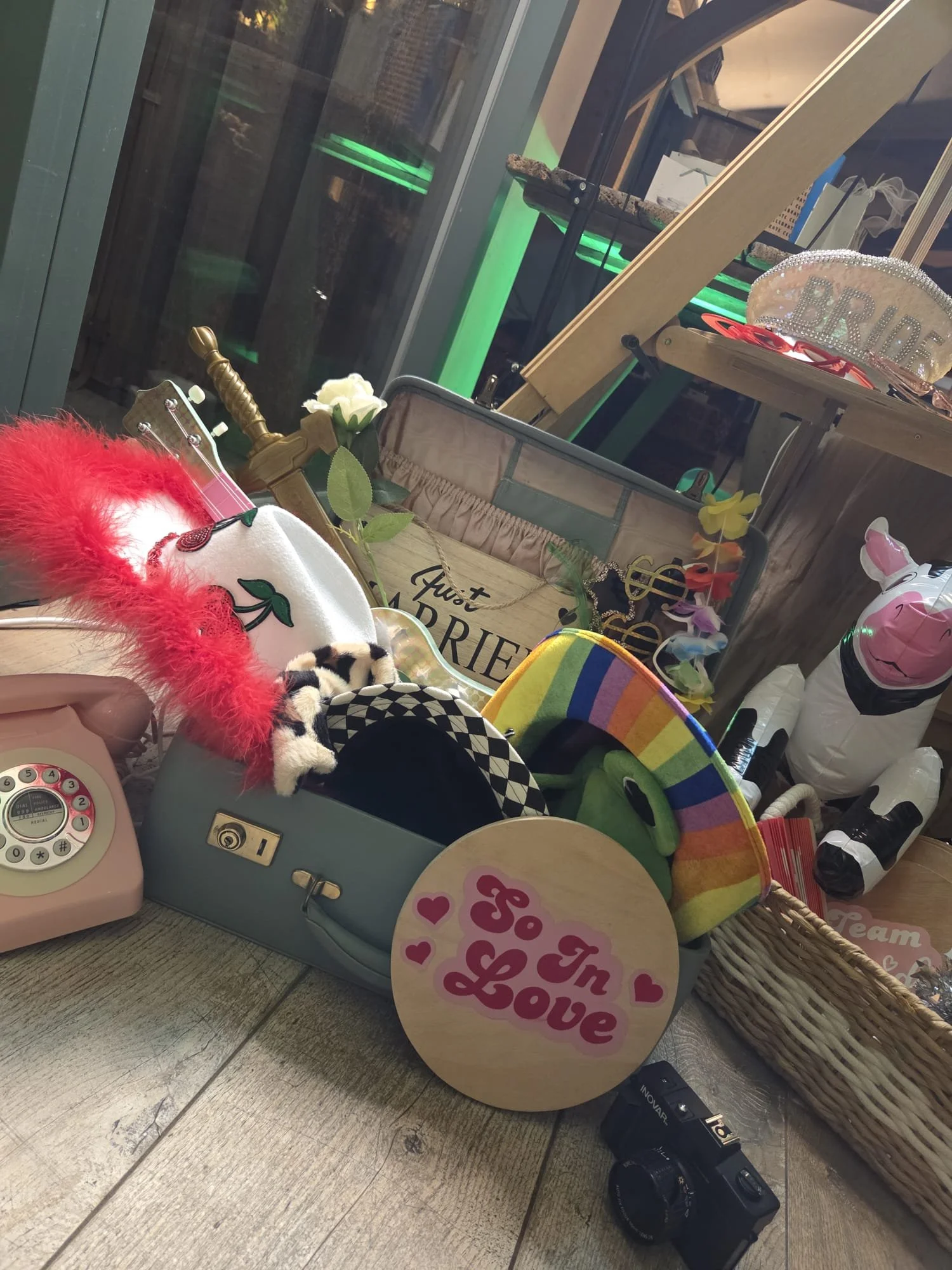 A basket filled with various birthday decorations and items, including a sign that reads 'Love', a pink and white unicorn figure, a rainbow-colored item, a stuffed animal, a telescope, and a wooden sign with 'Just a little bit of love'. In the background, there is a sign that says 'BRIDE' and a glass display with black and green lighting.
