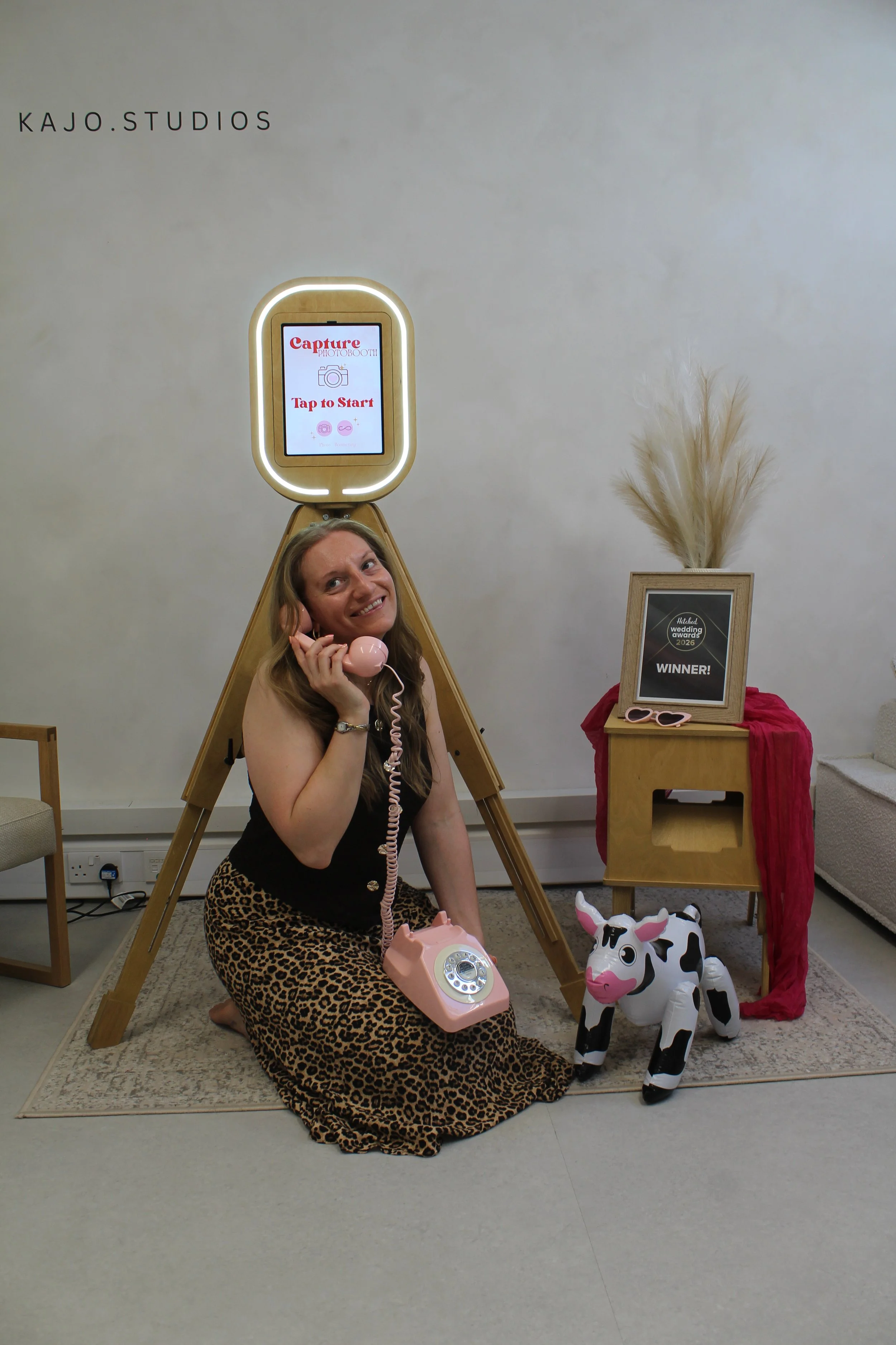 A woman sitting on the floor with a pink rotary phone, smiling. Behind her is a photo booth with a sign that says 'Tap to Start'. To her right is a decorated table with a framed 'WINNER!' sign, pampas grass, and pink sunglasses, with a cow-shaped inflatable toy on the floor.