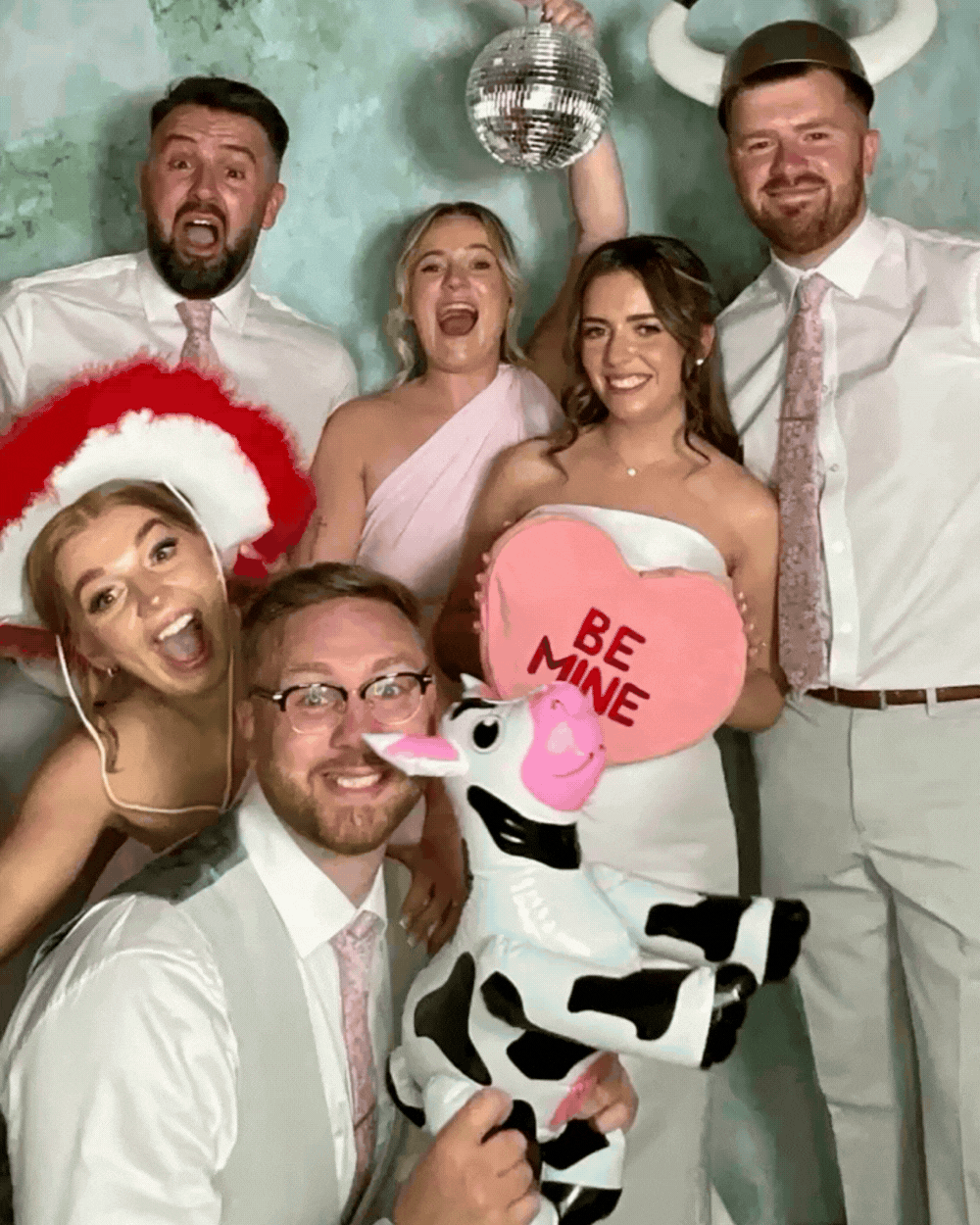 Group of six people at a celebration, some wearing wedding attire, with party props including a cow stuffed animal, a pink heart-shaped sign that says "Be Mine," and festive accessories. They are smiling and appear to be having fun.