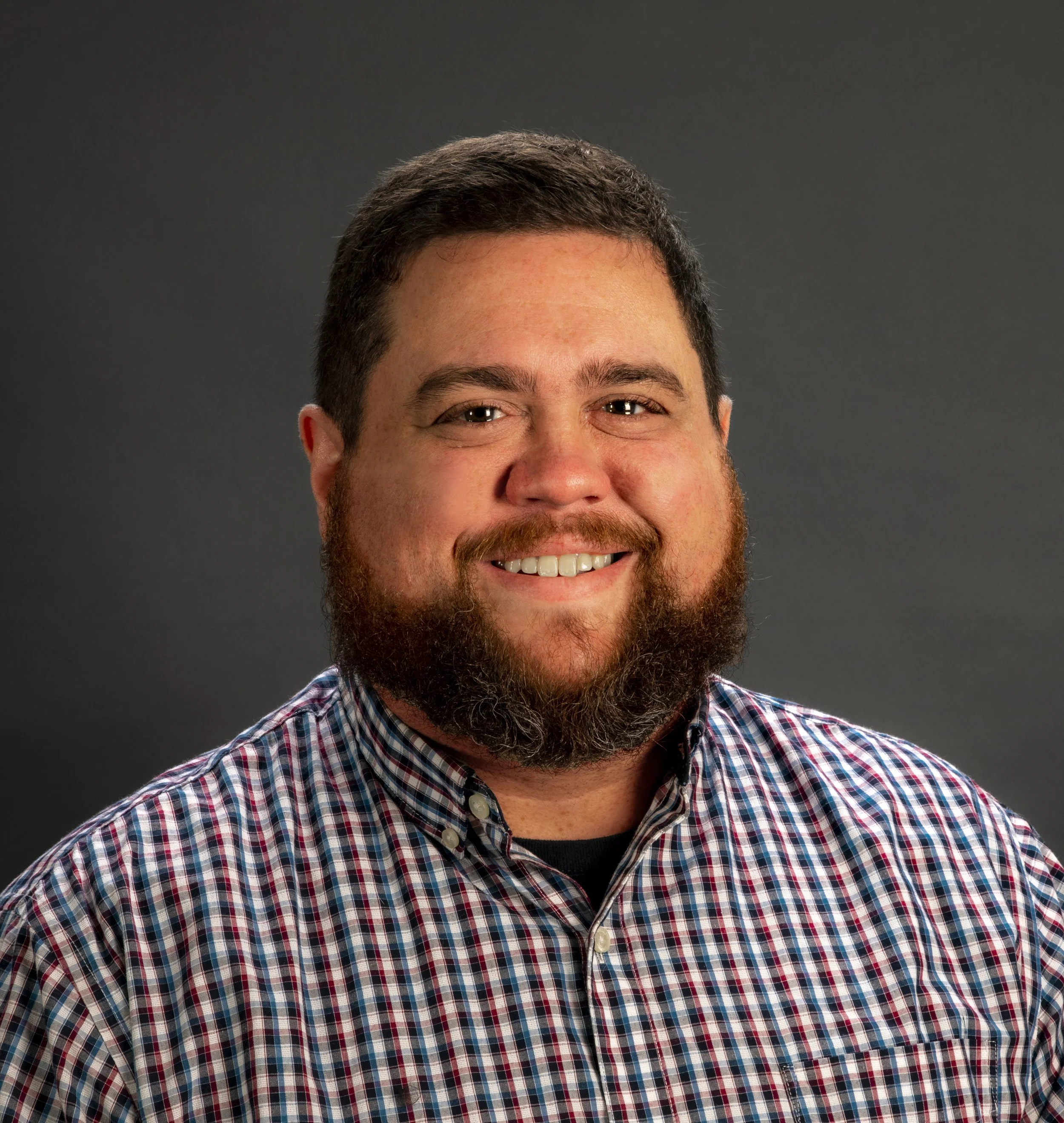 Headshot of a man with dark hair and a beard, smiling, wearing a plaid shirt, against a dark gray background.