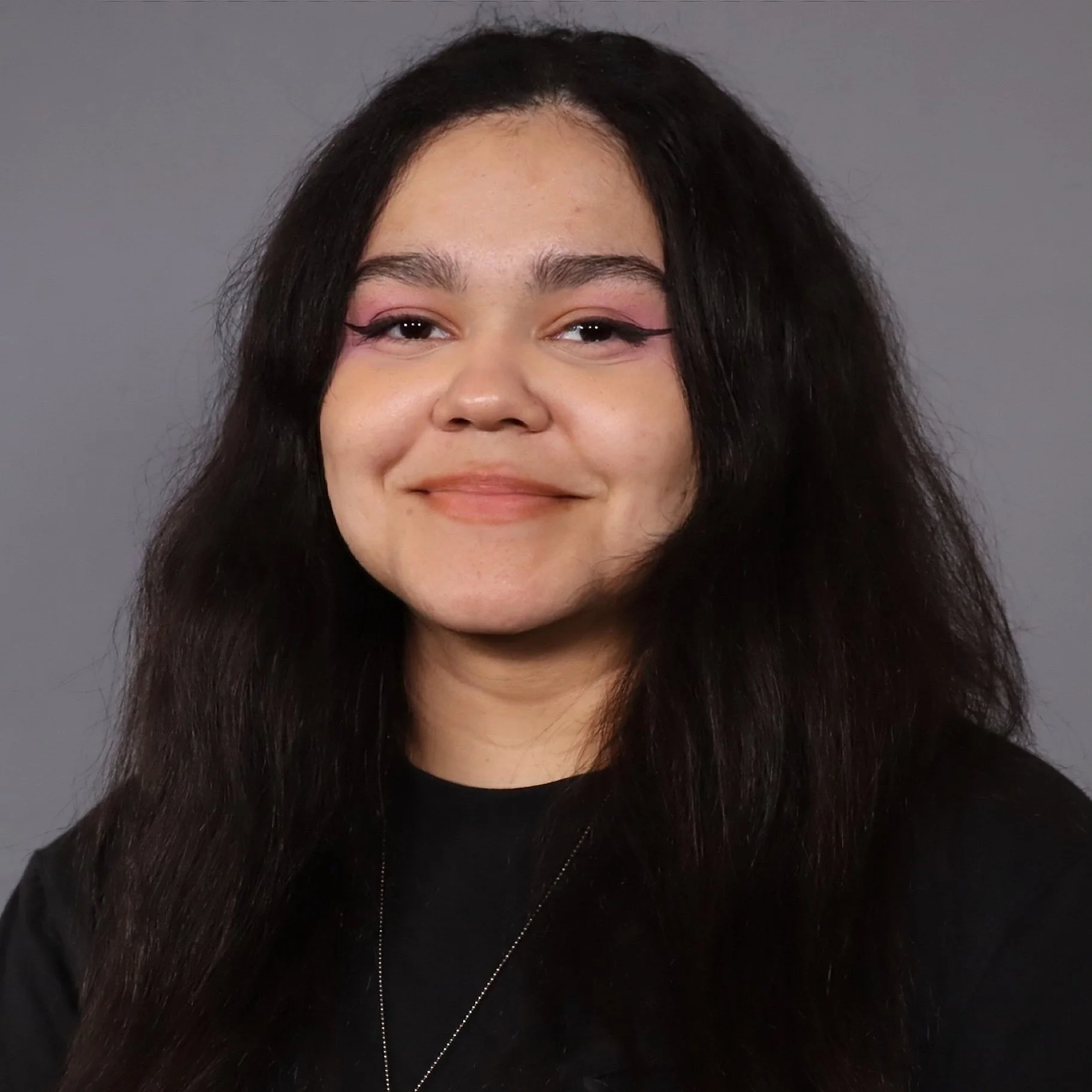 A young woman with long dark hair and light makeup, smiling at the camera against a gray background.