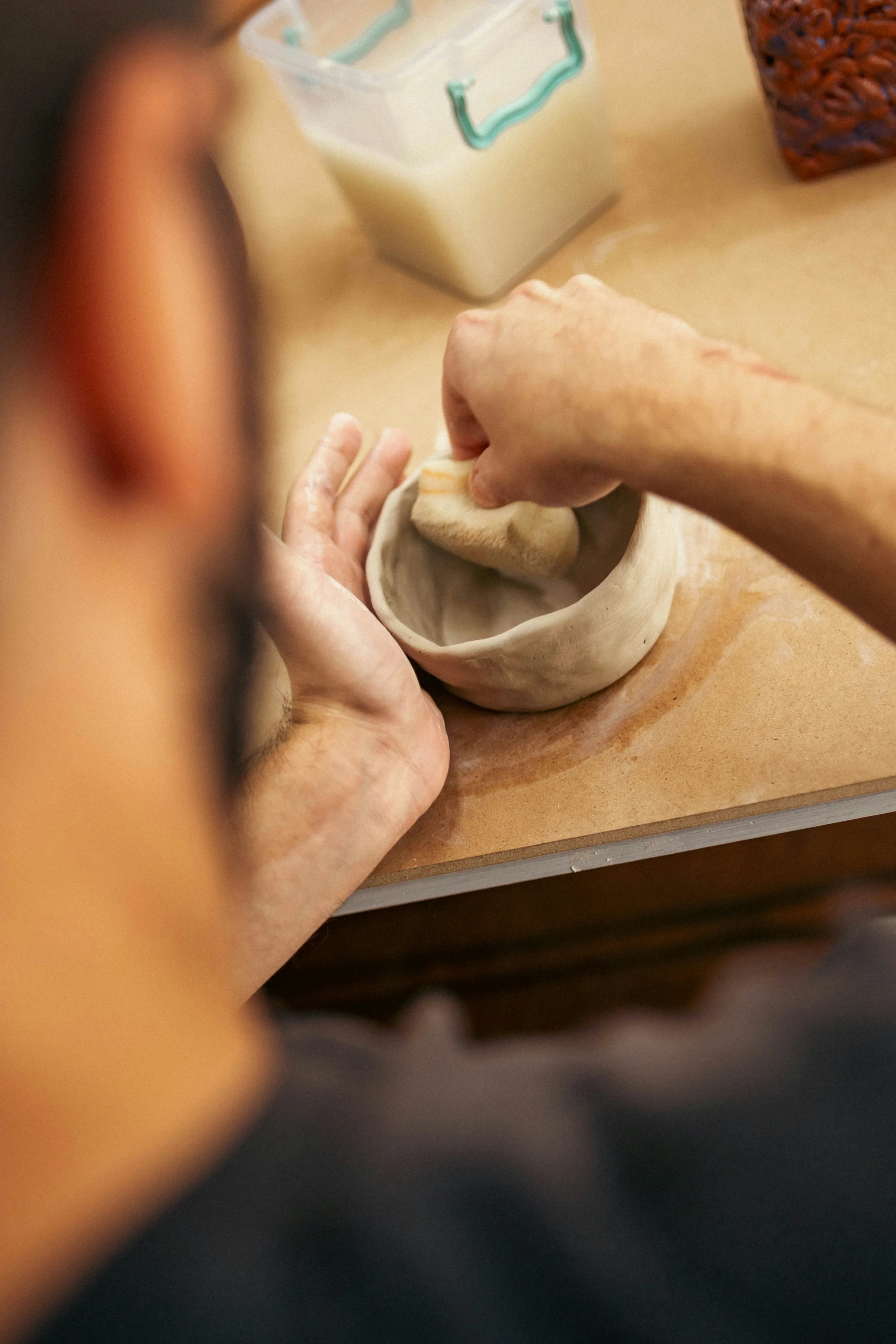 A person is working with clay, shaping it into a bowl or dish. Their hand is pressing into the clay, and some flour or powder is on their fingers. There are various objects on the table, including a container with a teal lid and a woven basket.