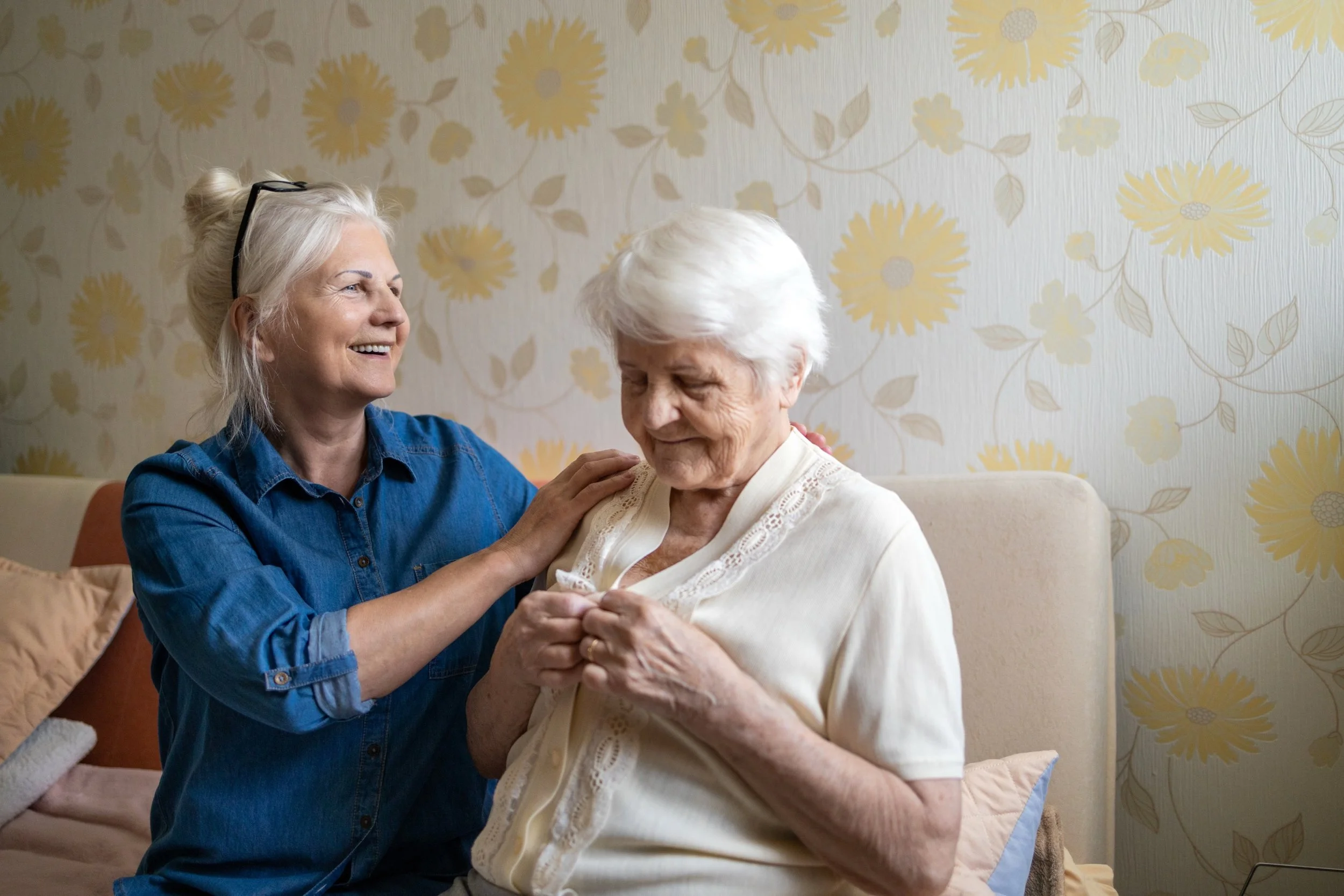 A woman with silver hair and glasses on her head smiling and holding hands with an elderly woman with white hair, who is also smiling and wearing a cream-colored blouse. They are sitting in a room with floral wallpaper.