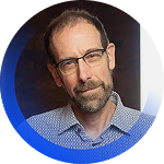 Headshot of a man with glasses, short dark hair, and a beard, wearing a blue checkered shirt, in front of a dark background with a circular blue border.