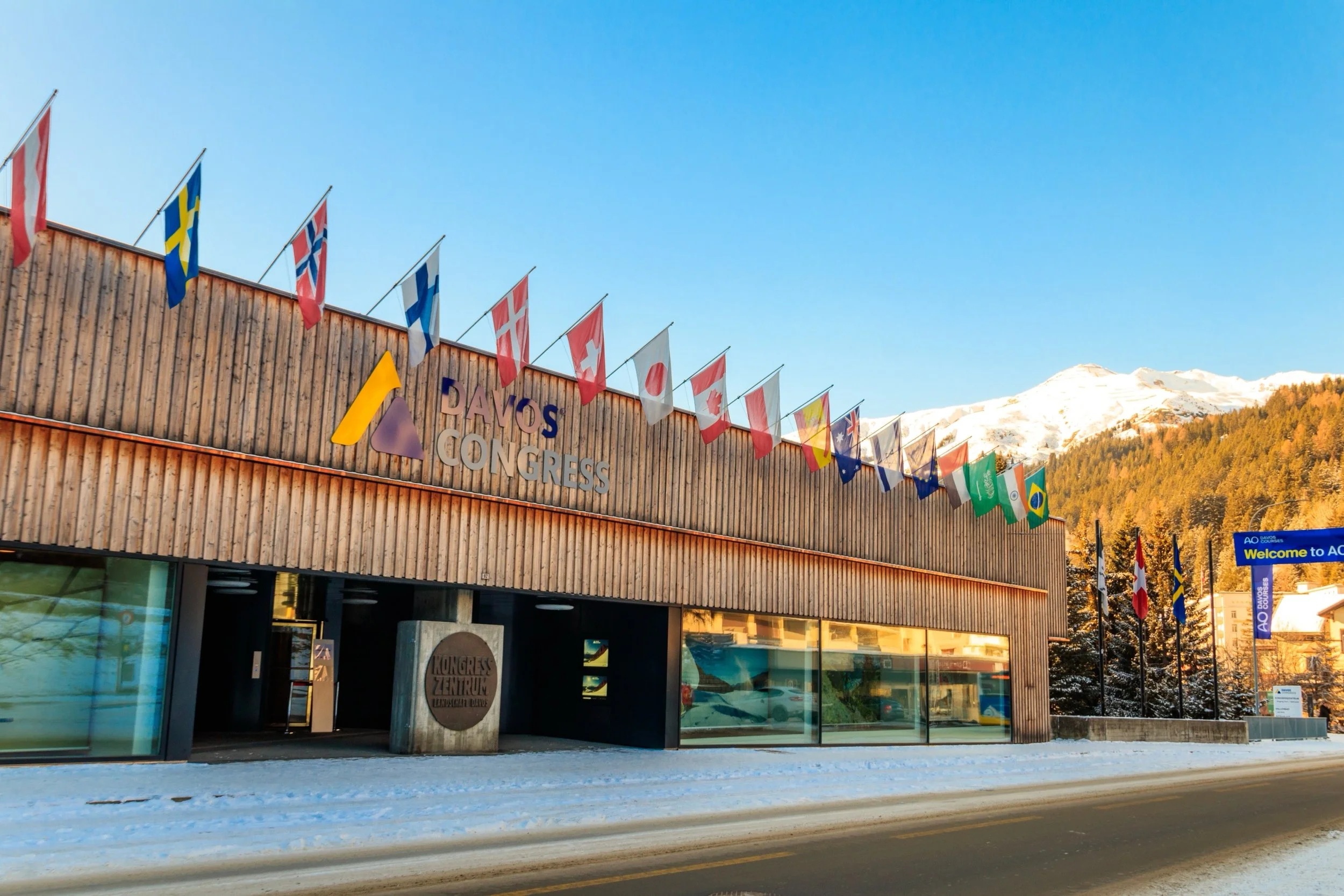 The Davos Congress center building with international flags, snowy mountains in the background, and snow on the ground.