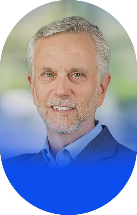 Headshot of a middle-aged man with gray hair, beard, wearing a blue shirt and blazer, smiling against a blurred outdoor background.