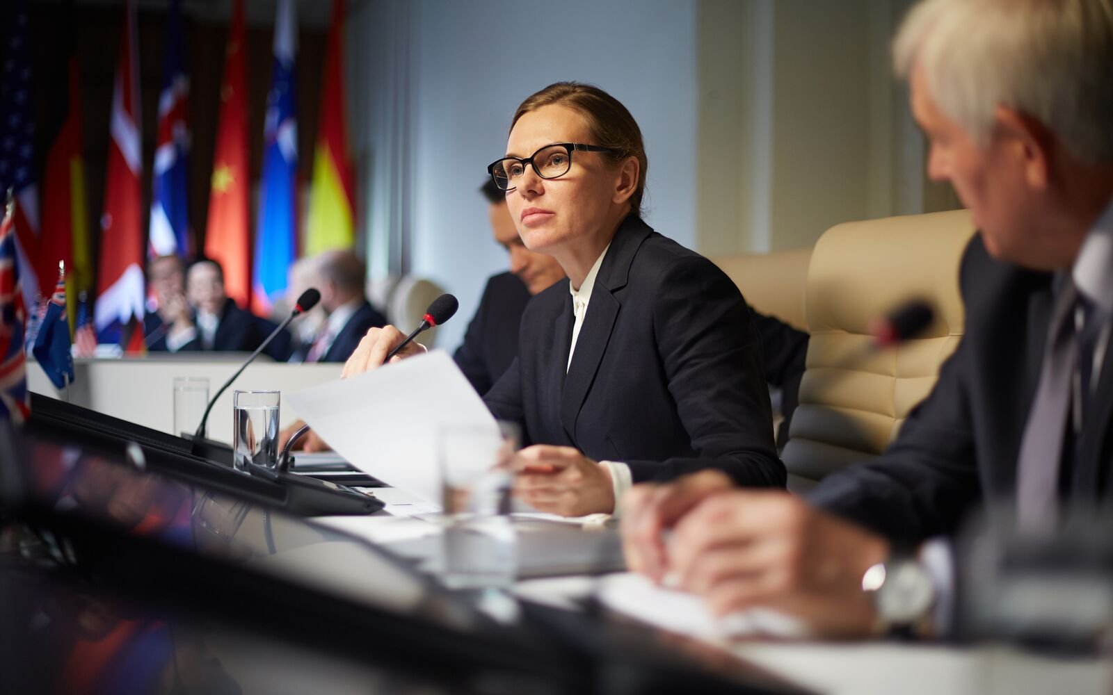 A woman in a black suit and glasses sitting at a conference table with multiple flags in the background, speaking into a microphone during an international meeting.