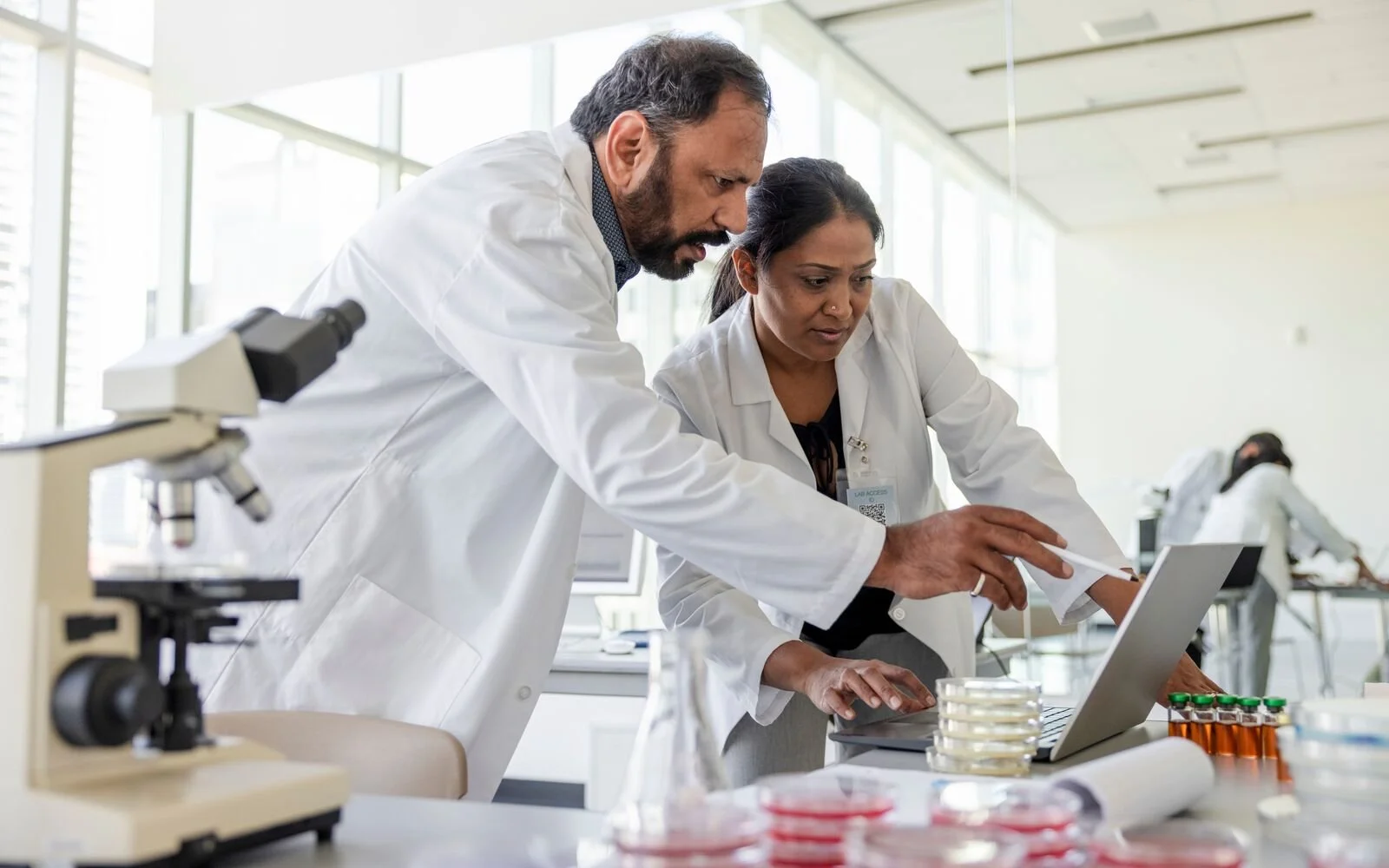 Two scientists, a man and a woman, in lab coats working together on a laptop in a modern laboratory with microscopes and laboratory equipment visible.
