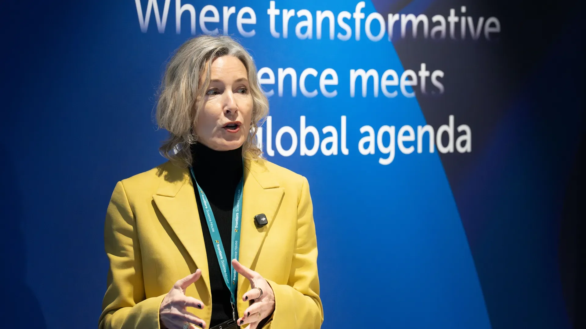A woman with shoulder-length blonde hair wearing a yellow blazer and black turtleneck speaks at a conference or event. She has a blue lanyard around her neck and a small microphone clipped to her blazer. Behind her, there is a large sign with the text "Where transformative science meets global agenda" against a blue background.