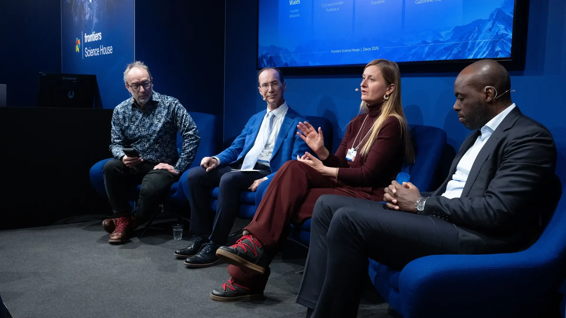 Four people seated on blue chairs participating in a panel discussion at a conference with a blue backdrop that reads 'Frontiers Science House.' The woman on the right is speaking, and the others are listening.