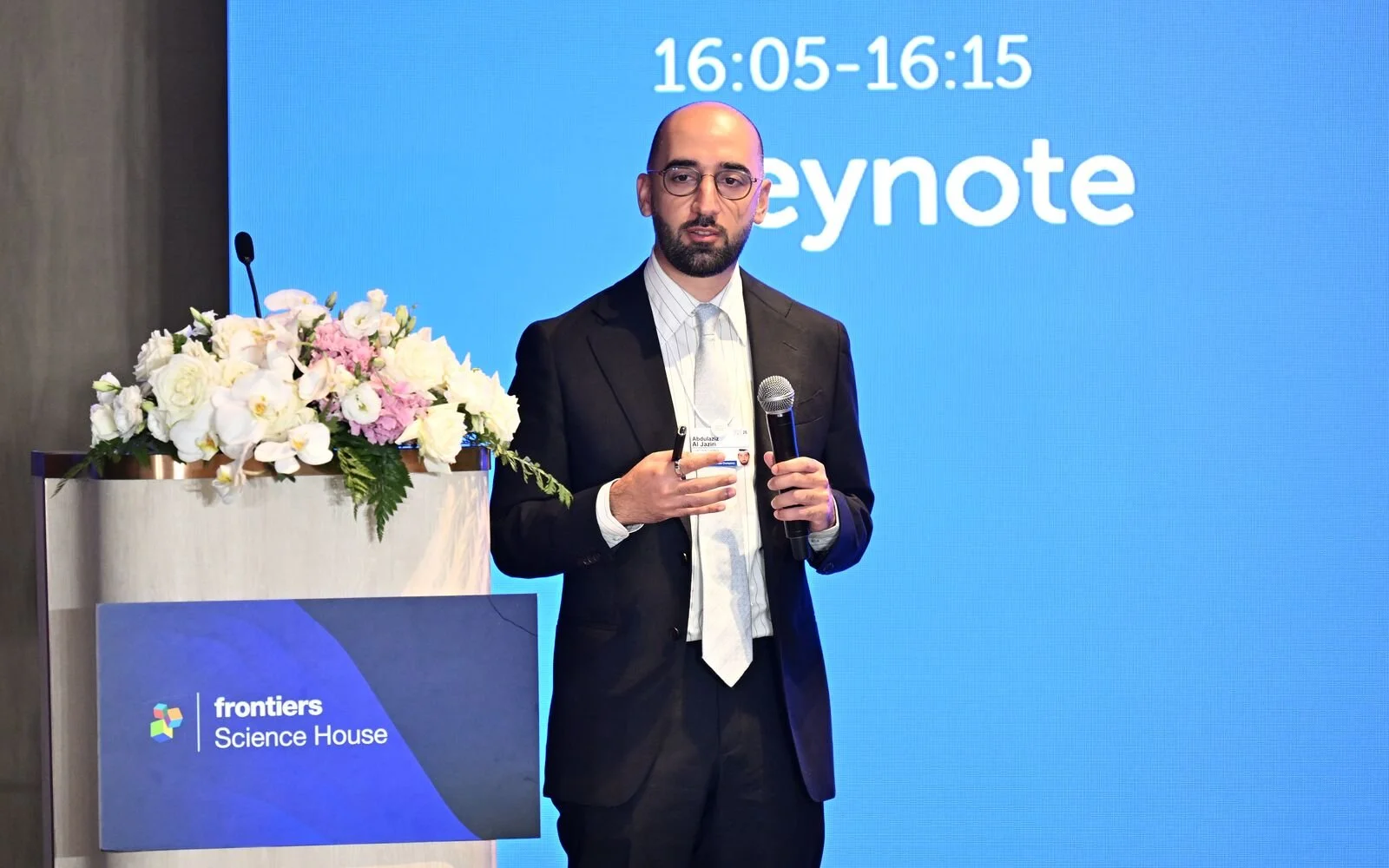 A man in a suit holding a microphone and a tablet, standing on stage at a conference with the logo of Frontiers Science House and a floral arrangement. Behind him, a blue screen displays the words 'keynote' and a time range.