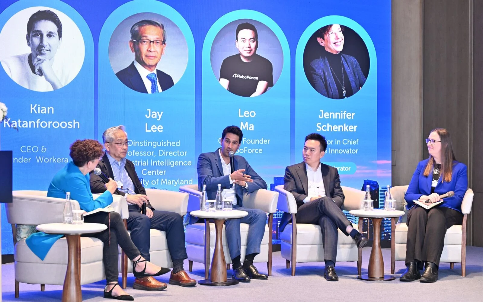 Panel discussion with five people seated on stage in front of a large blue digital screen displaying profile photos and names of four speakers: Kian Katanforoosh, Jay Lee, Leo Ma, and Jennifer Schenker. The panelists are engaged in conversation, with one holding a microphone and two taking notes.