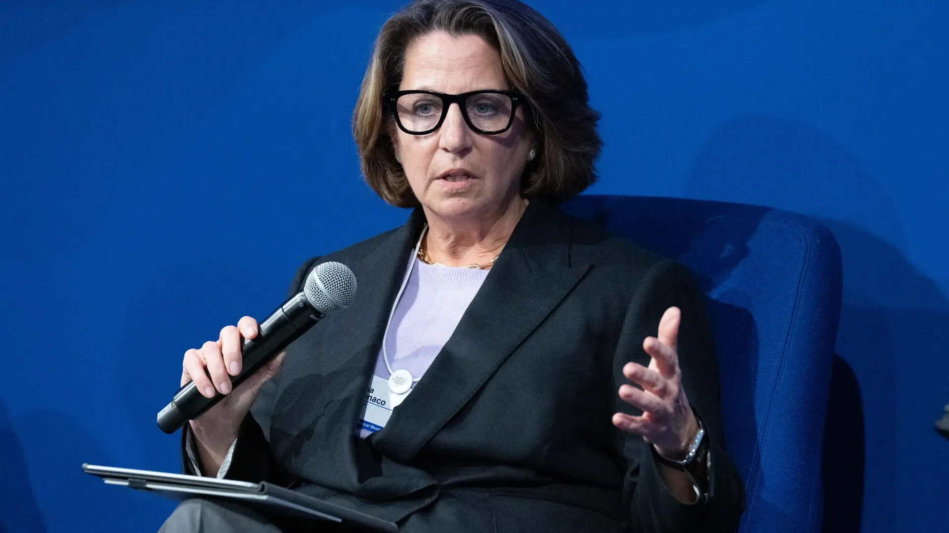 A woman with shoulder-length brown hair, black glasses, and wearing a black blazer speaking into a microphone while sitting in a blue chair against a blue background.