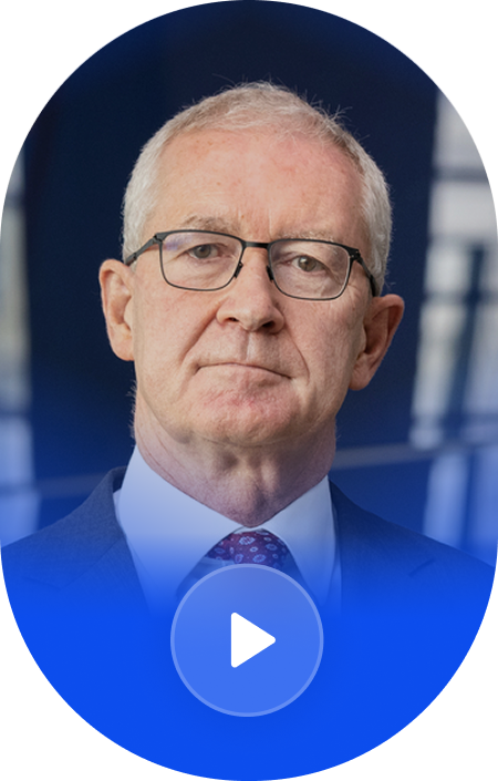 A close-up portrait of an elderly man with glasses, short white hair, wearing a suit and tie, in front of a blurry blue background.