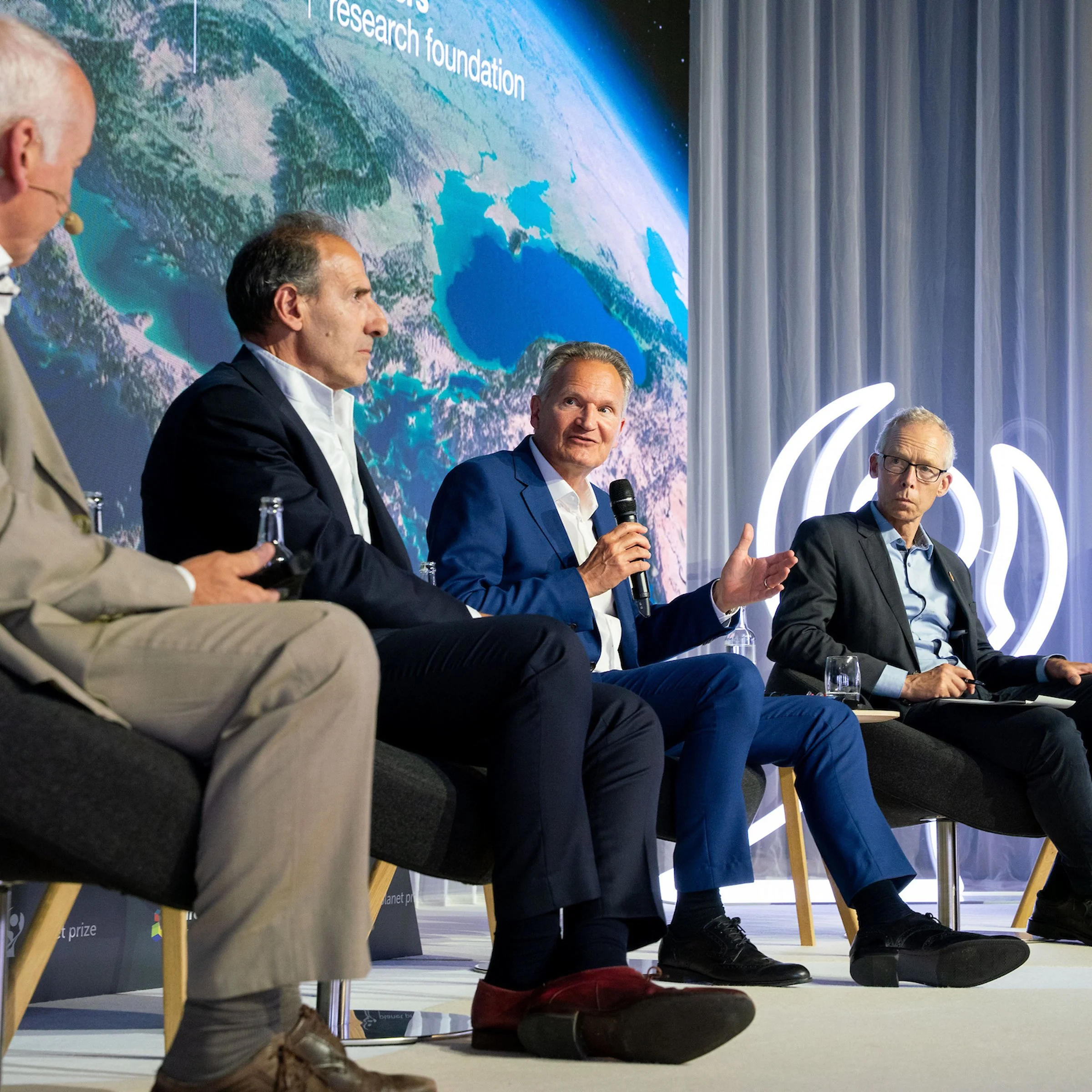 Panel of five men sitting on stage during a conference or panel discussion. One man is speaking into a microphone, and a large digital screen behind them displays a satellite image of Earth with some text.