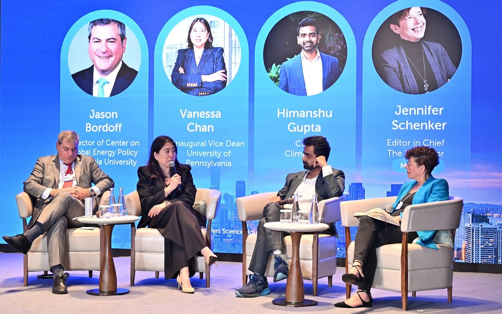 A panel discussion features four professionals seated on stage with a large blue screen behind them displaying their names, titles, and photos. The screen shows Jason Bordoff, Vanessa Chan, Himanshu Gupta, and Jennifer Schenker. Three individuals are actively engaged in conversation, while one appears to be listening or taking notes.