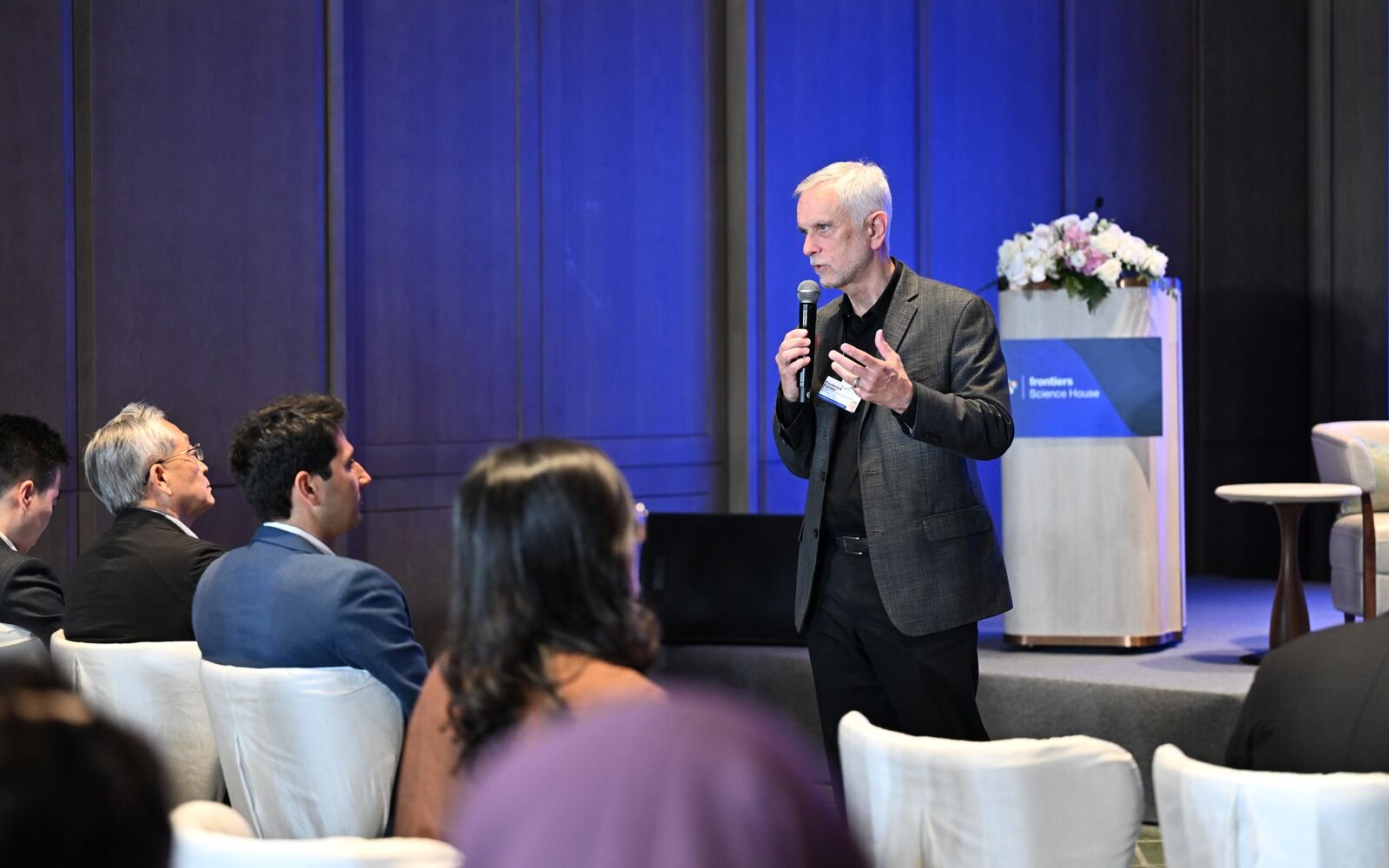 A man with white hair and a dark blazer is speaking into a microphone while gesturing with his hand during a presentation at a conference. Audience members are seated and listening attentively.
