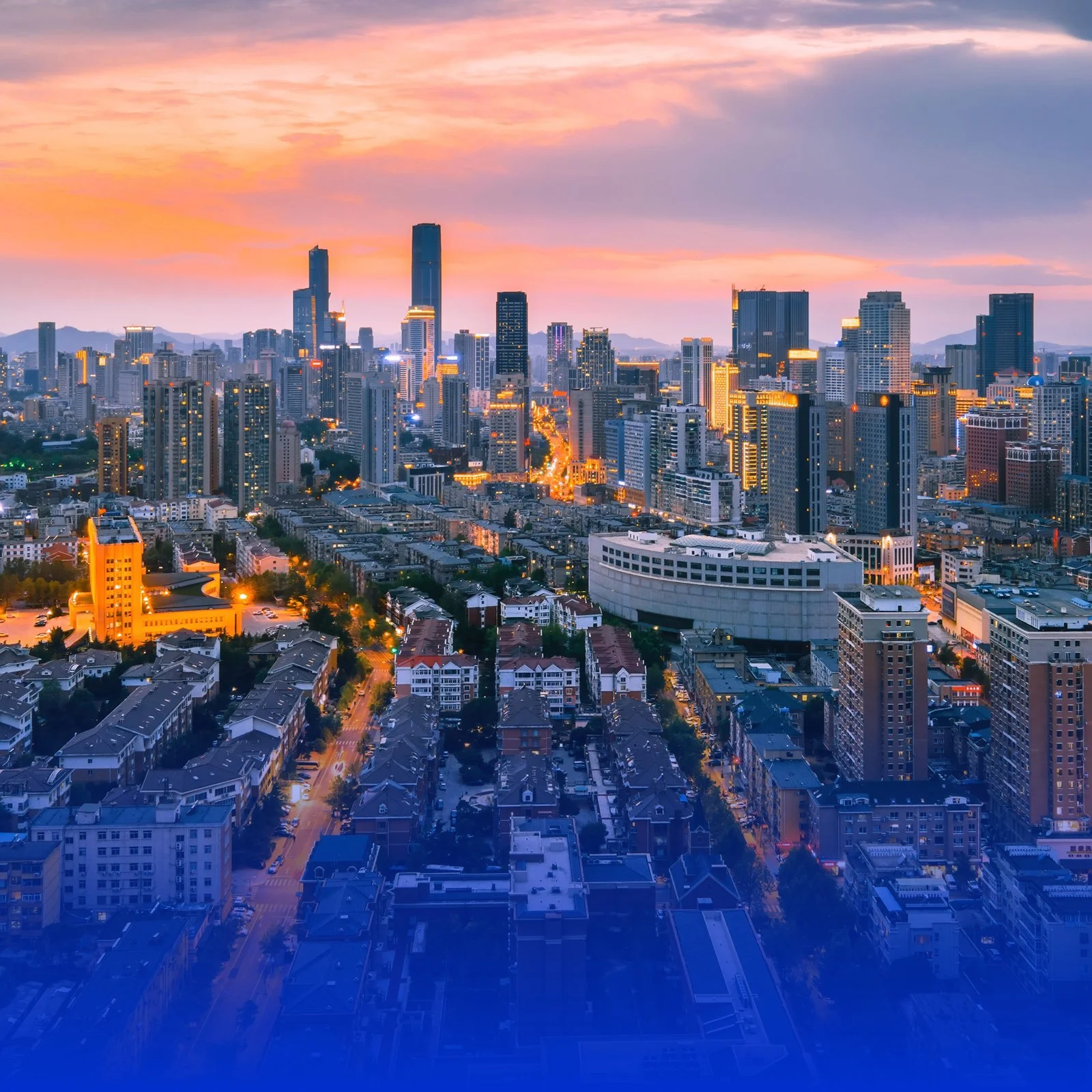 A city skyline at sunset with tall skyscrapers and residential buildings, illuminated by streetlights and building lights, against a sky with pink and orange hues.