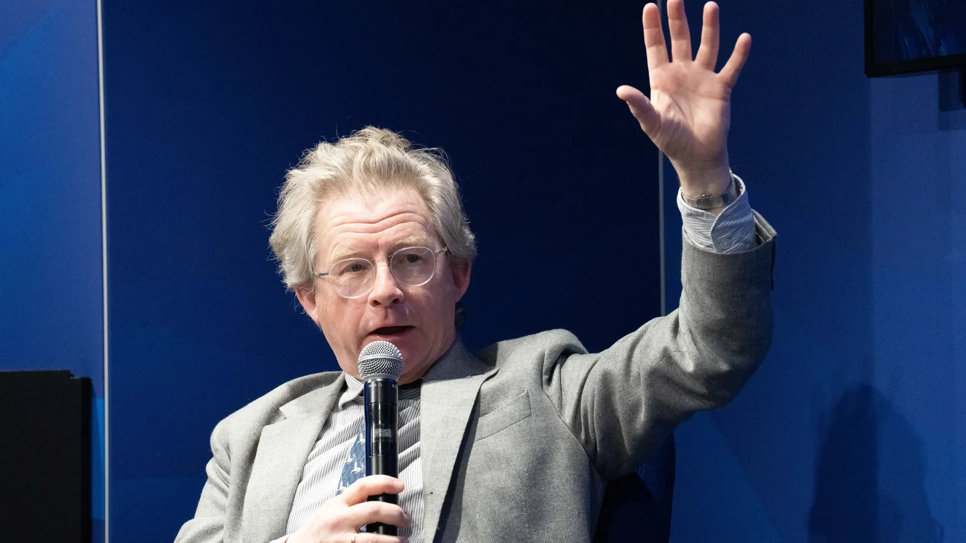 An older man with glasses and gray hair, wearing a gray suit, speaking into a microphone and raising his right hand.
