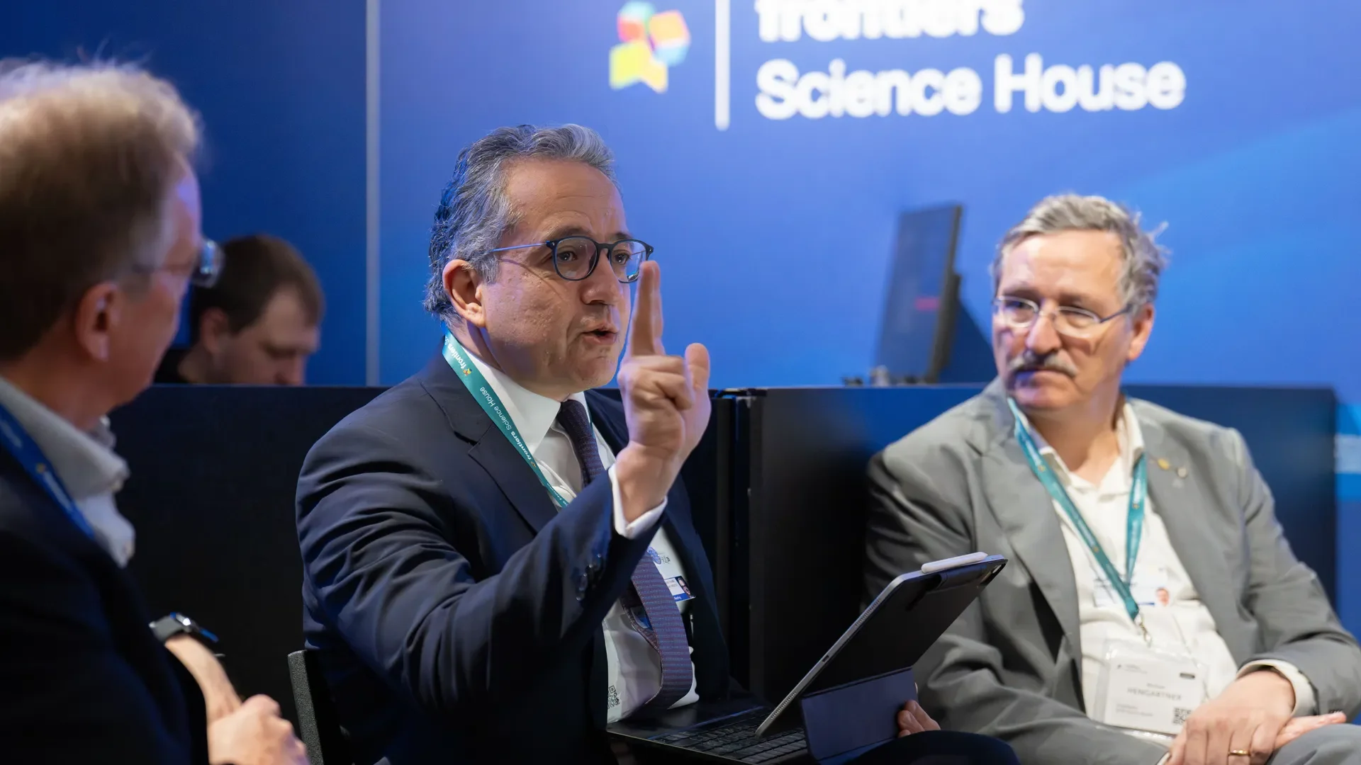 People sitting in a panel discussion at a science event, with a man in a suit and glasses speaking and gesturing with his finger, and a blue backdrop with the words 'Science House' visible.