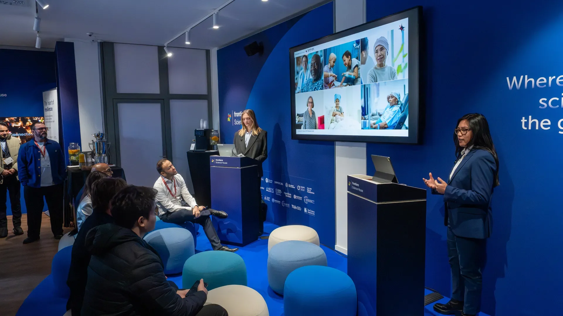 A woman and a woman speaking at a conference or presentation in a room with blue walls and a large screen displaying images of diverse healthcare professionals, including patients and medical staff. Audience members are seated and listening, with some taking notes or using laptops.