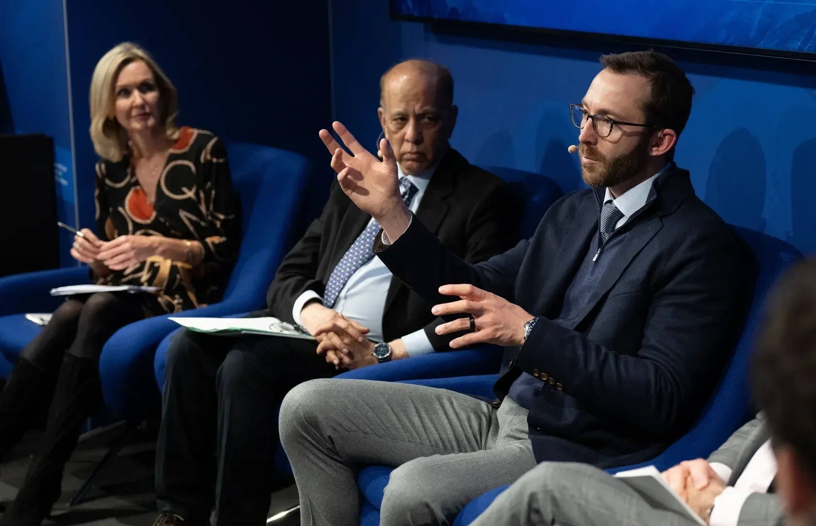 Three people sit in a row of blue chairs during a conference or panel discussion. The middle person is an older man with a serious expression, dressed in a black suit and tie. The person on the right is a man with glasses and a beard, wearing a dark jacket and a tie, gesturing with his hand. The woman on the left has blonde hair, wears a patterned dress, and holds some papers and a pen.