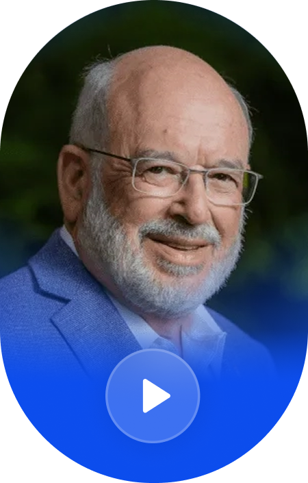 Close-up photo of an older man with glasses, white beard, smiling, wearing a blue blazer and white shirt, outdoors with blurred green background.