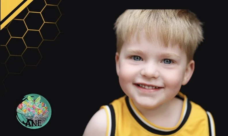 Smiling young boy with blond hair and blue eyes wearing a yellow sports jersey, against a black background with a honeycomb pattern and a small logo in the lower left corner.