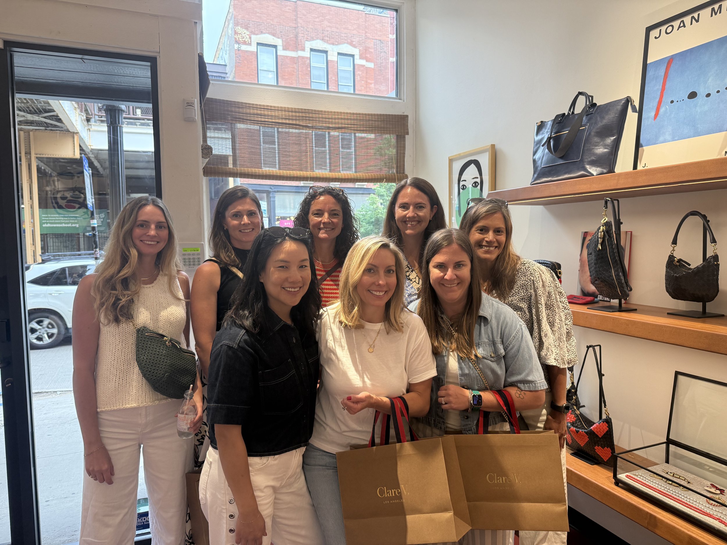 Group of women standing inside a store, smiling at the camera. The store has shelves with handbags and accessories, and a window showing buildings outside.