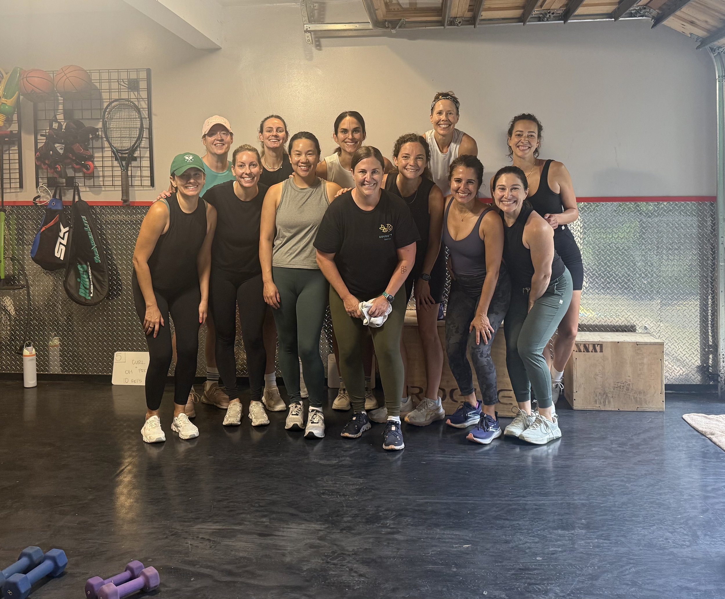 A group of fifteen women posing together in a gym, smiling. They are dressed in workout clothes, with some wearing tank tops and leggings. The gym has a black rubber floor and gym equipment on the wall, including tennis rackets, basketballs, and tennis balls. Dumbbells are in the foreground, and a small sign is on the wall behind them.