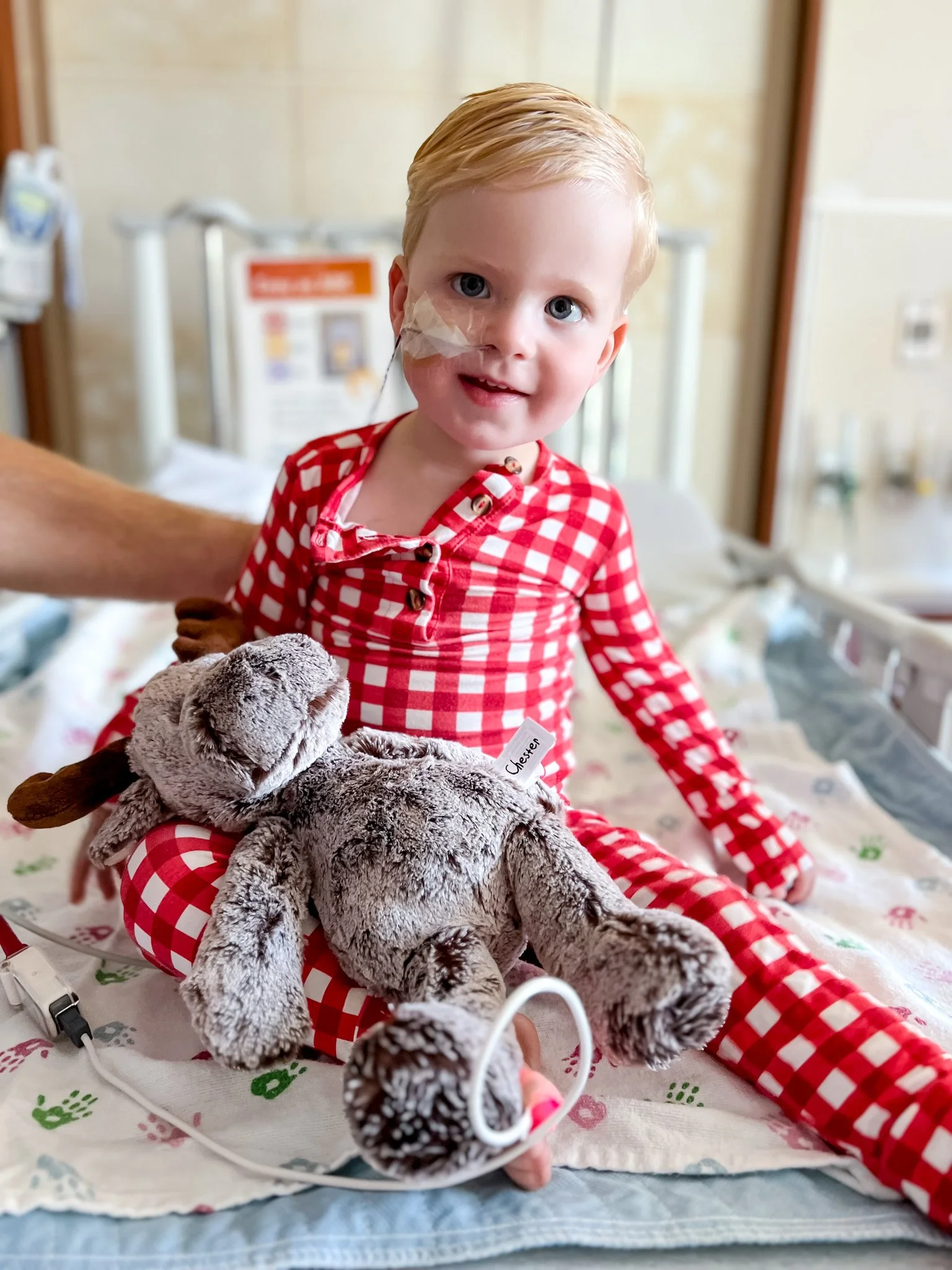 A young child with a bandage on their face, lying in a hospital bed, smiling while holding a stuffed animal, surrounded by medical equipment.