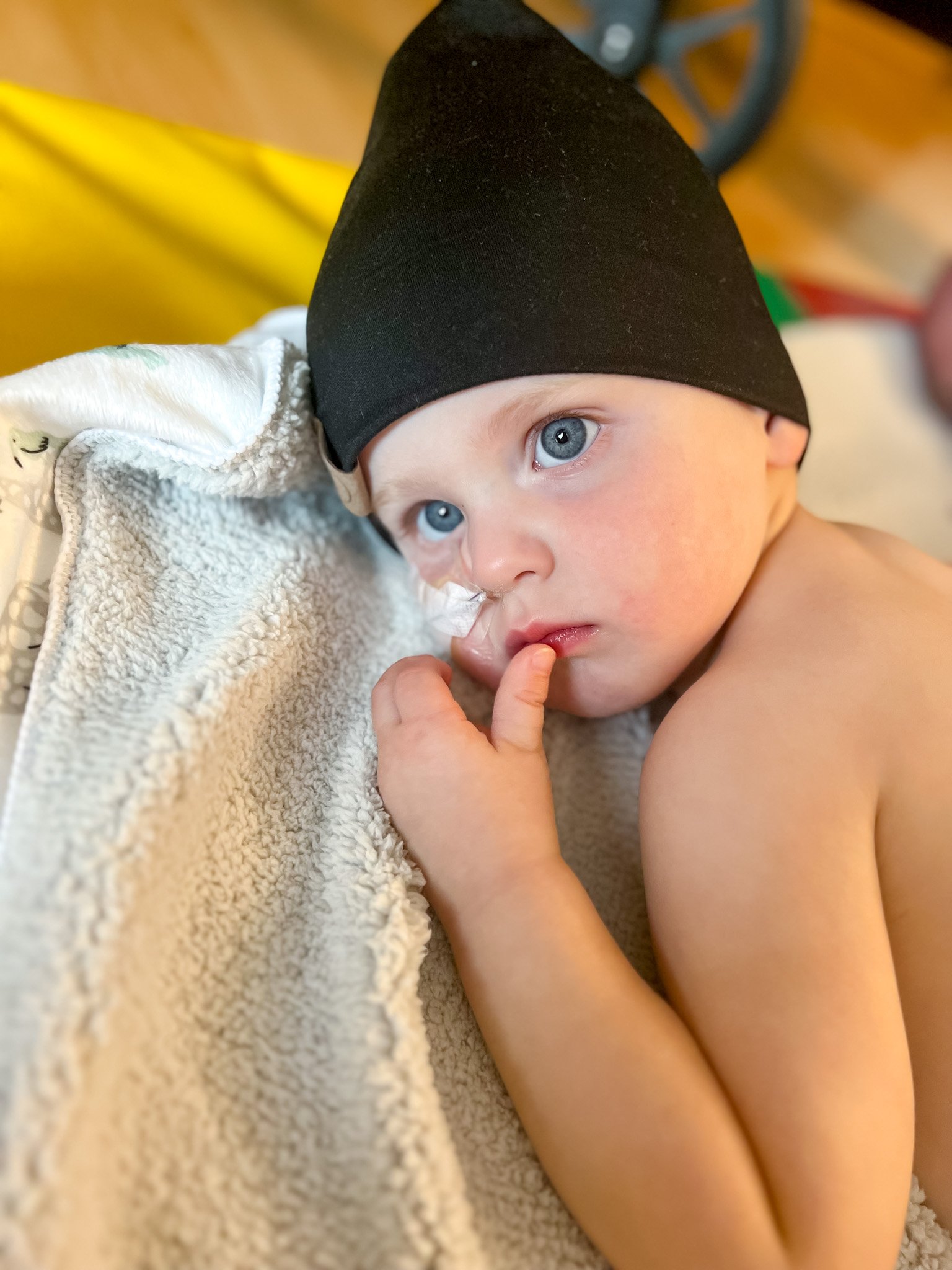 A young child with blue eyes, wearing a black cap, lying on a beige blanket, touching their lips with their finger.
