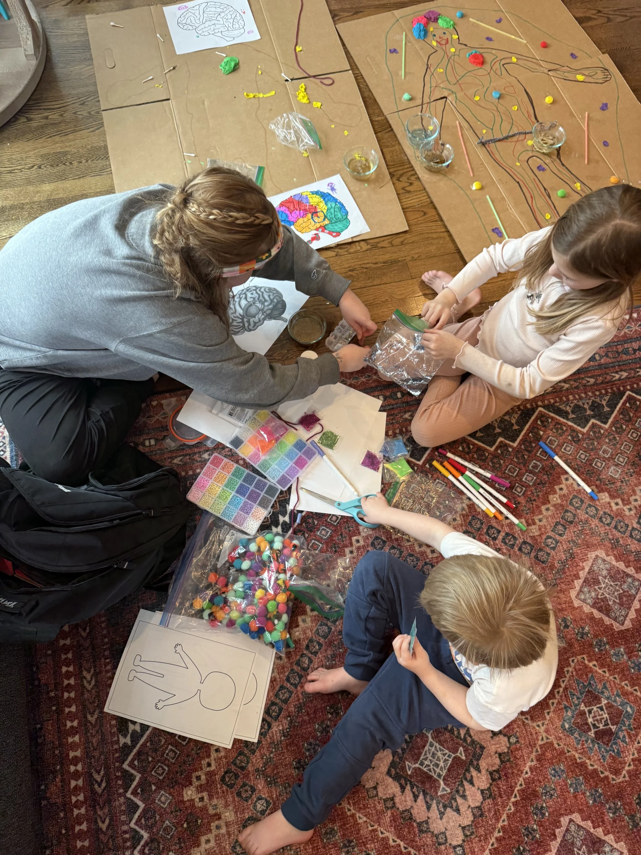 Kids creating a colorful origami or craft project on a patterned rug, surrounded by black and white drawings, colorful pom-poms, scissors, and craft supplies.