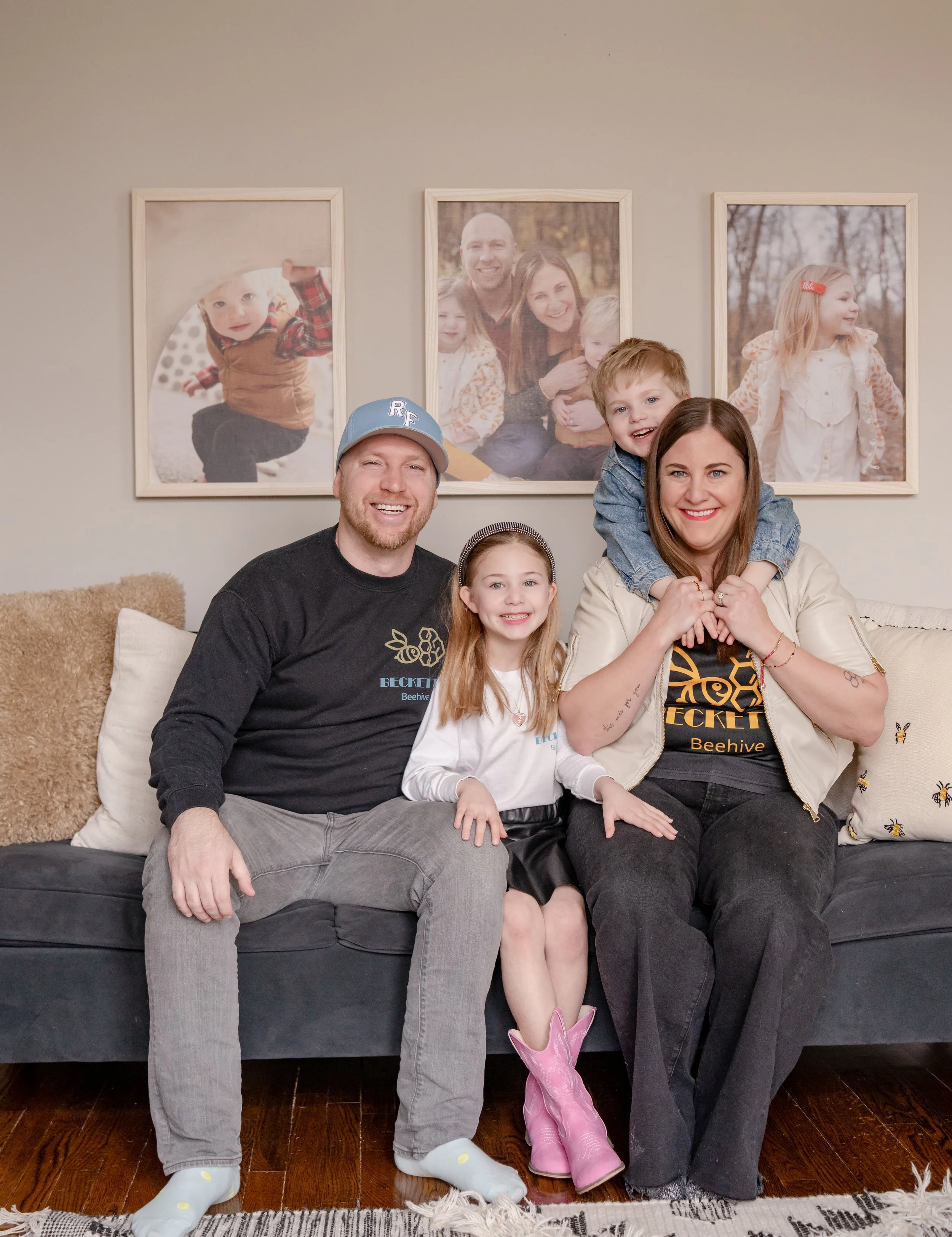 A family of four sitting on a black sofa in a living room. The man and woman are smiling, and the two children, a girl and a boy, are also happy. The girl is wearing pink cowboy boots and the boy is wearing pink rain boots. The background features three framed family photos on a gray wall.