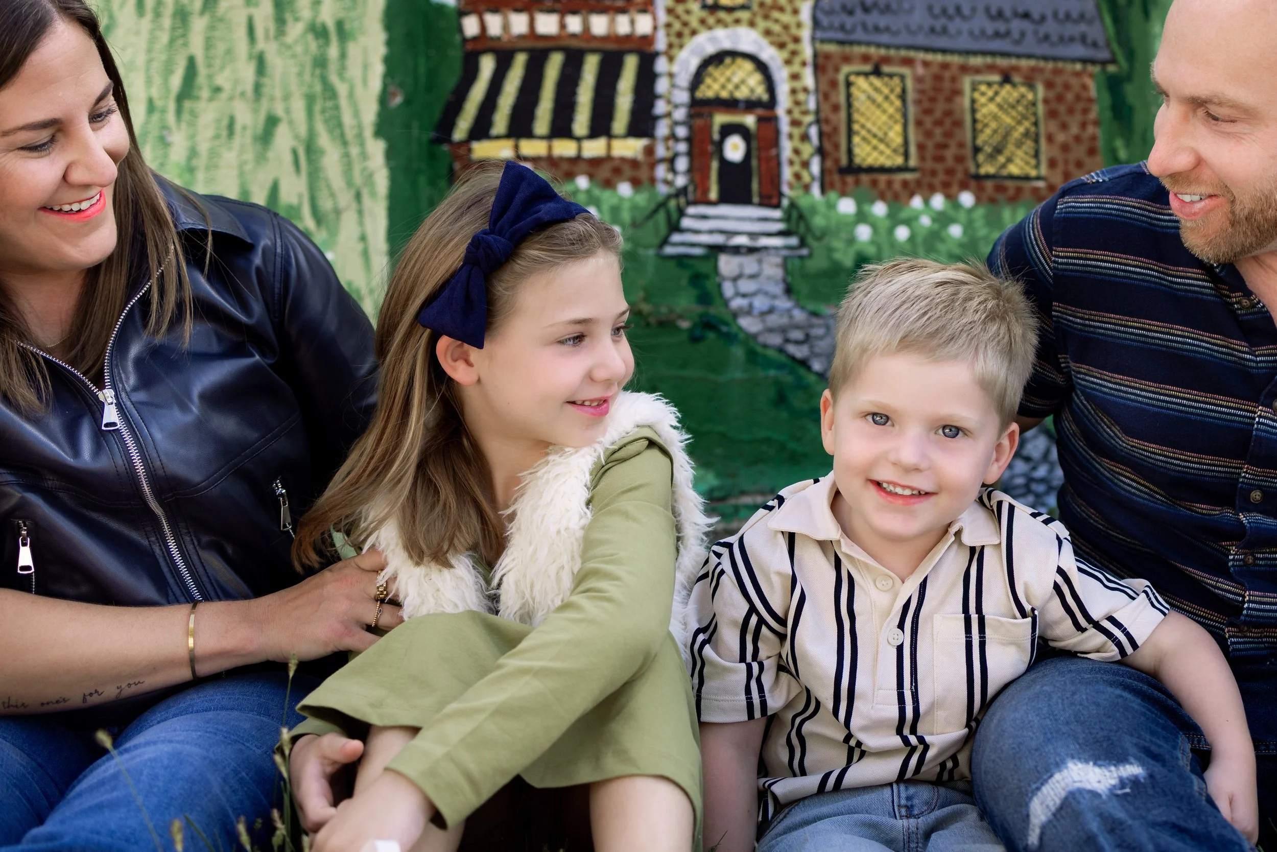 A group of smiling people including three adults and two children sitting together outdoors with a colorful brick house painted mural in the background.