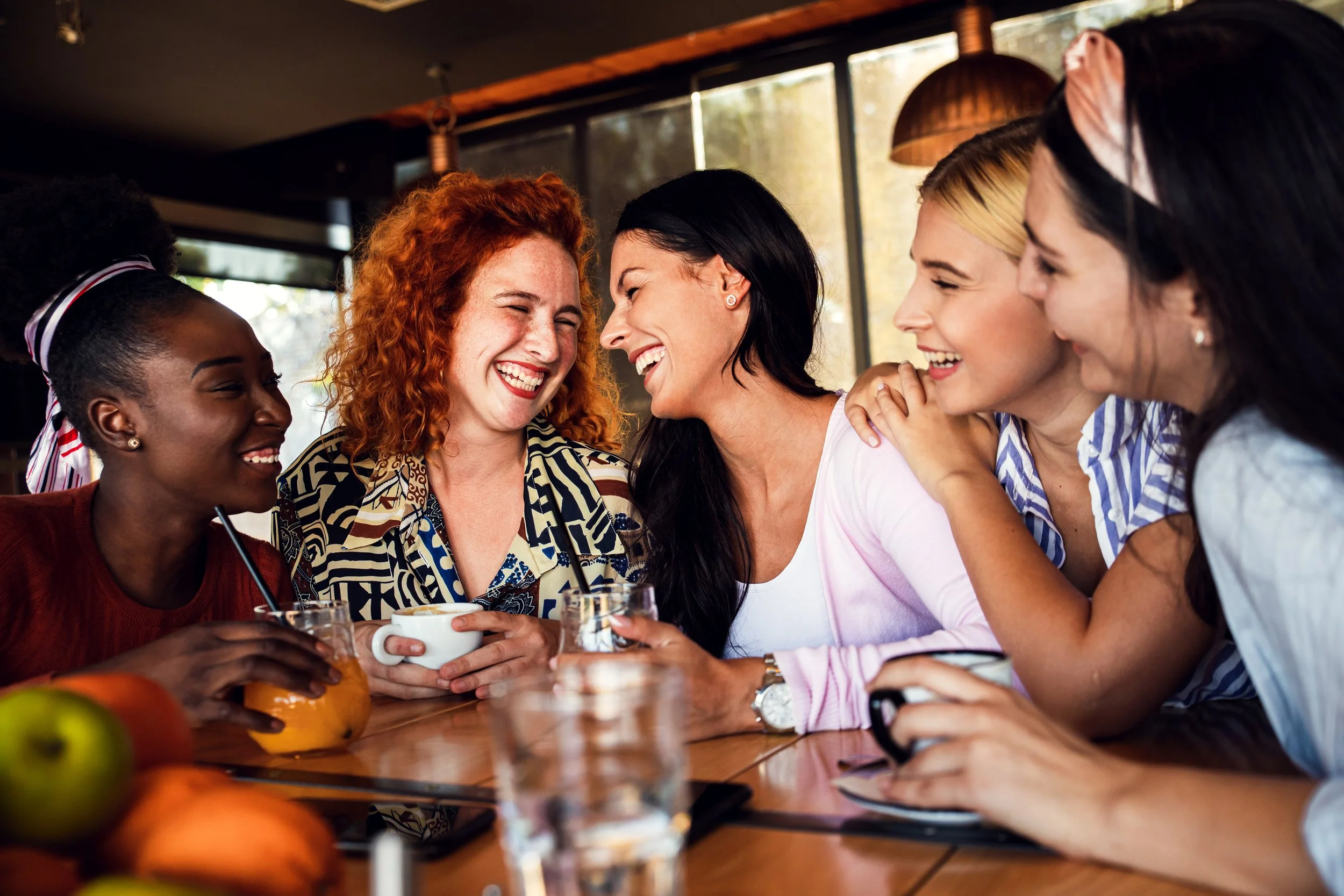 Group of women laughing and enjoying drinks at a social gathering in a cozy cafe or restaurant.