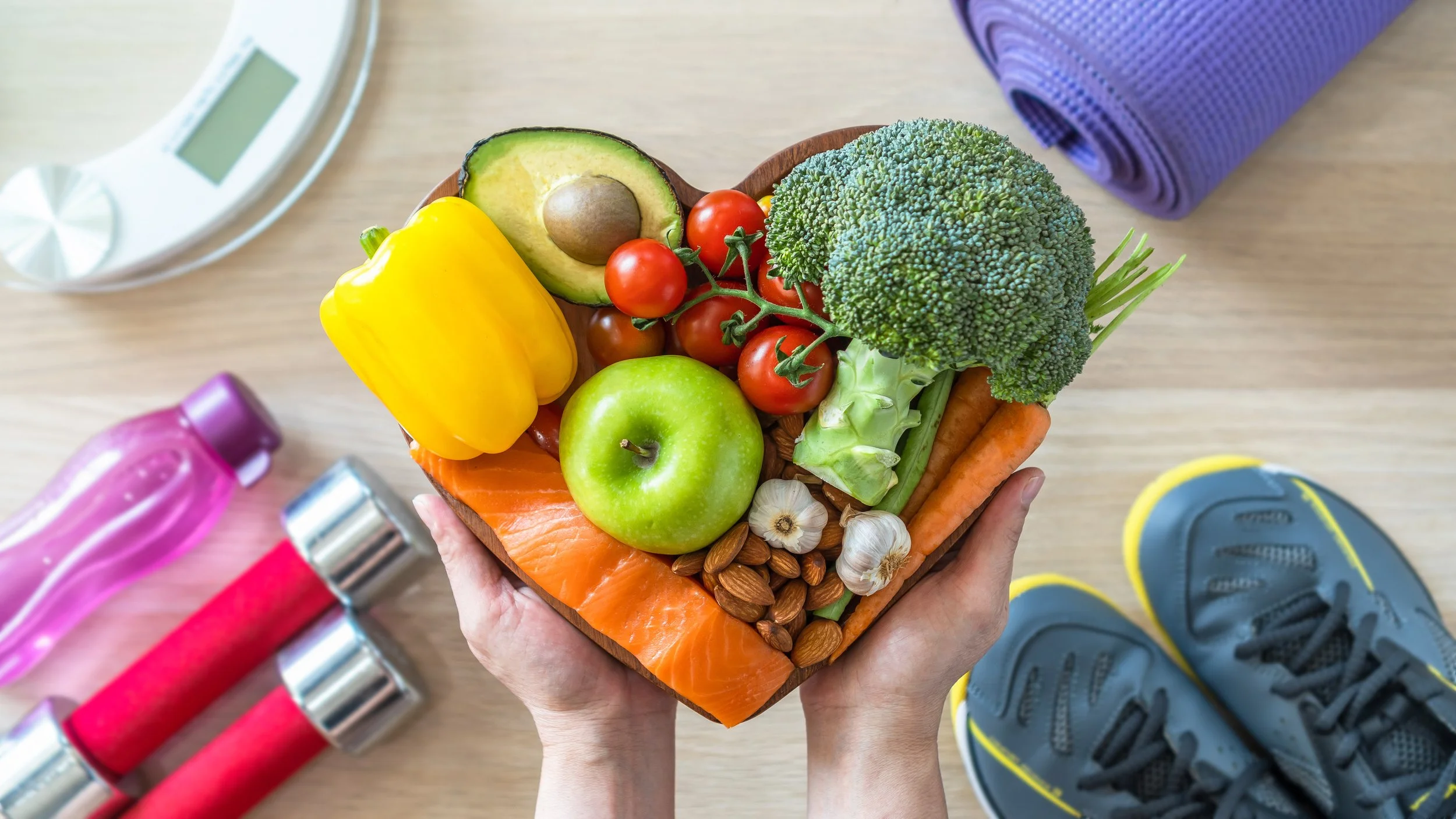 A heart-shaped wooden bowl filled with various fresh vegetables and fruits, including cherry tomatoes, broccoli, avocado, green apple, yellow bell pepper, garlic, and salmon, held by a person standing on a workout mat with sneakers nearby, surrounded by fitness equipment.