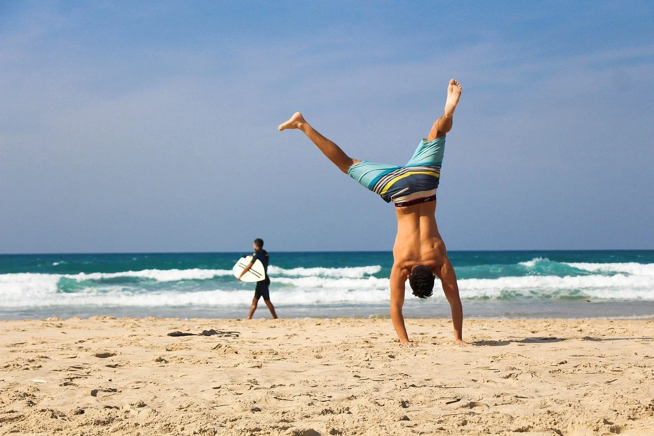 A man doing a handstand on the sandy beach near the ocean with a surfer walking in the background.