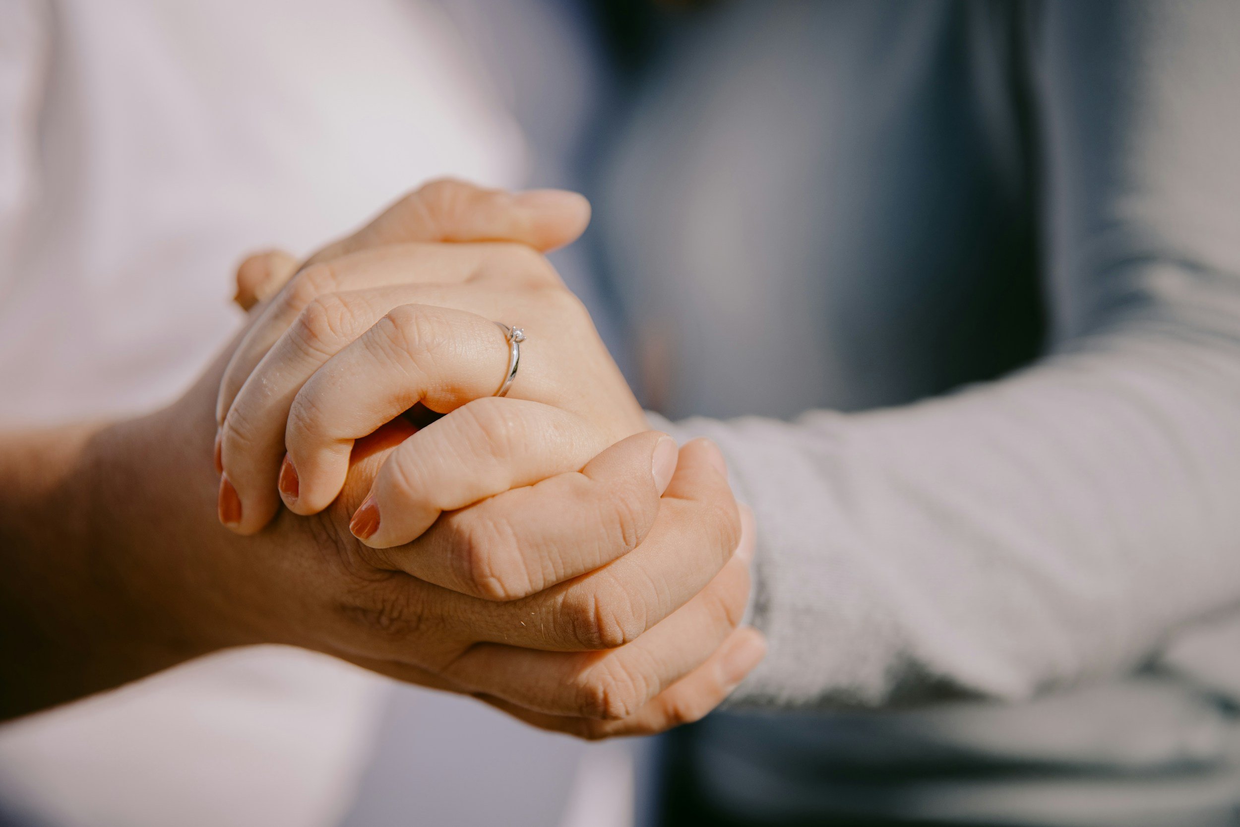 Close-up of two hands gently holding each other, one with a wedding or engagement ring, against a blurred background.