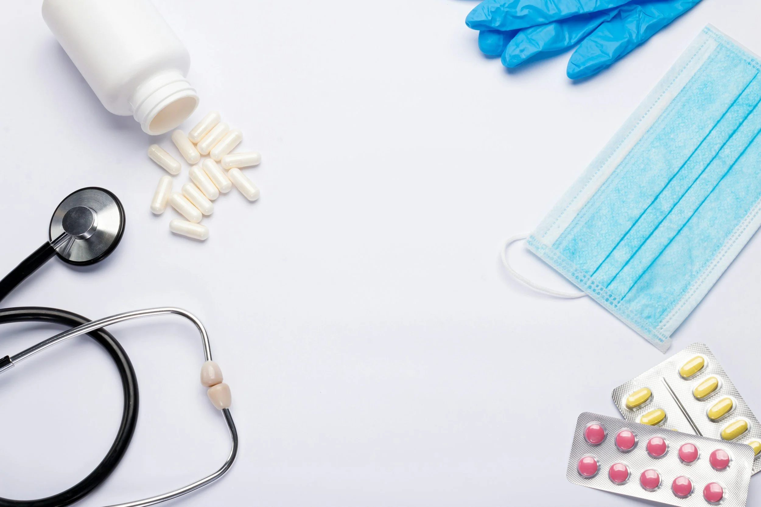 An arrangement of medical items on a white background, including a bottle of pills with some tablets spilled, a stethoscope, a pair of blue medical gloves, a blue surgical mask, and blister packs of pink and yellow capsules.