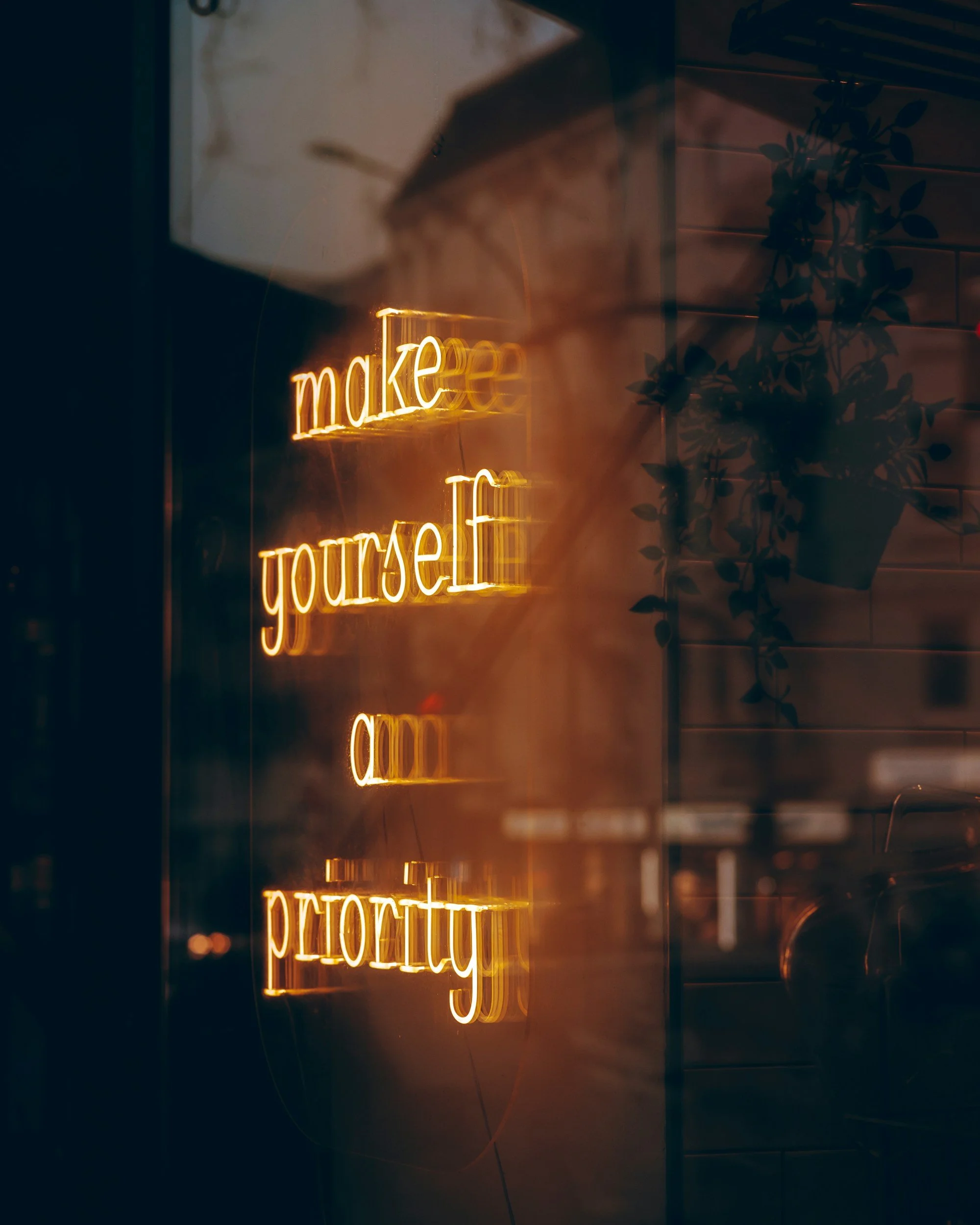 A neon sign with the words 'make yourself' in warm yellow-orange light, reflected on a glass surface next to a leafy plant.