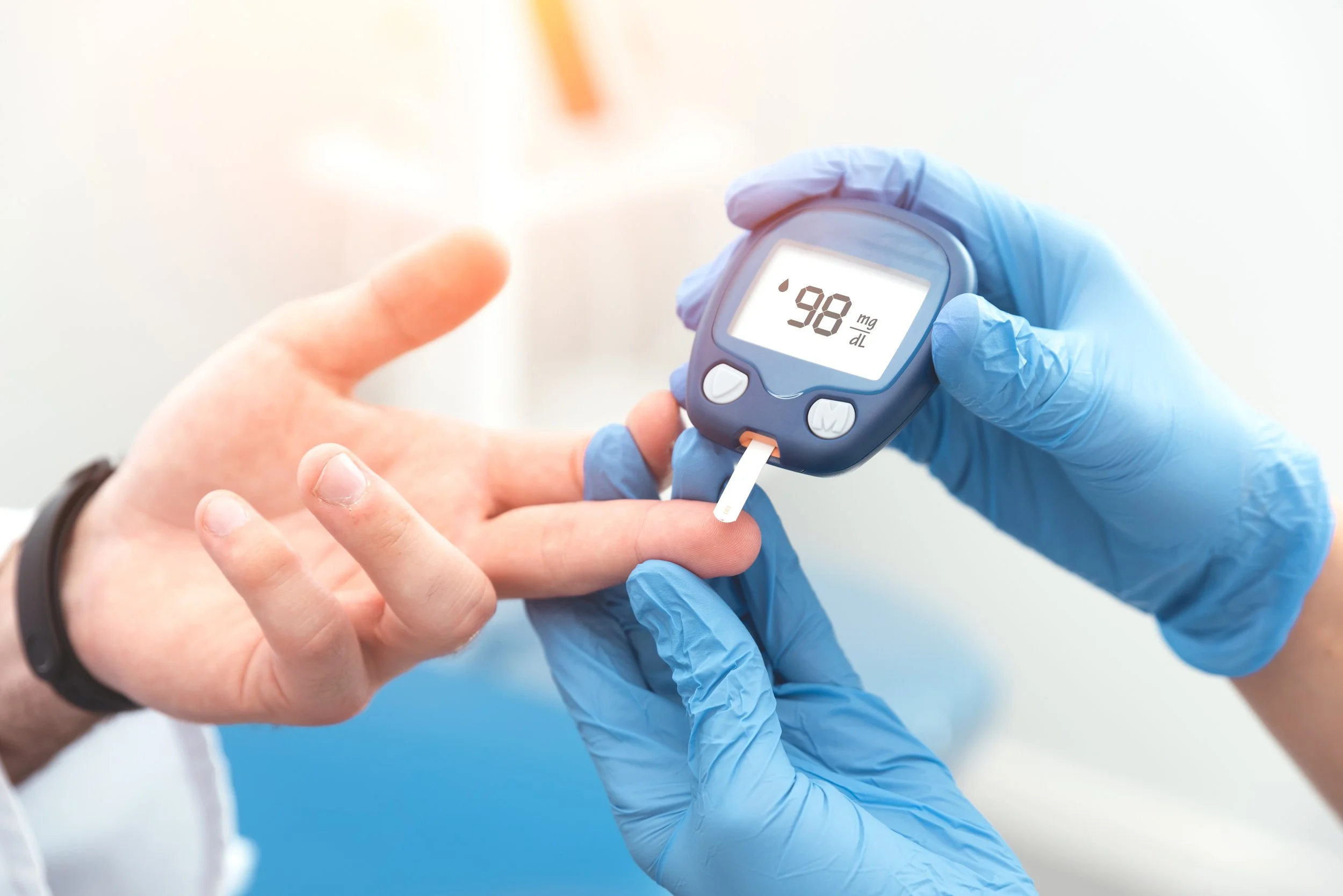A healthcare professional using a glucometer to check blood sugar levels of a patient. The glucometer screen displays a reading of 98 mg/dL. The professional is wearing blue gloves and the patient's hand is visible.