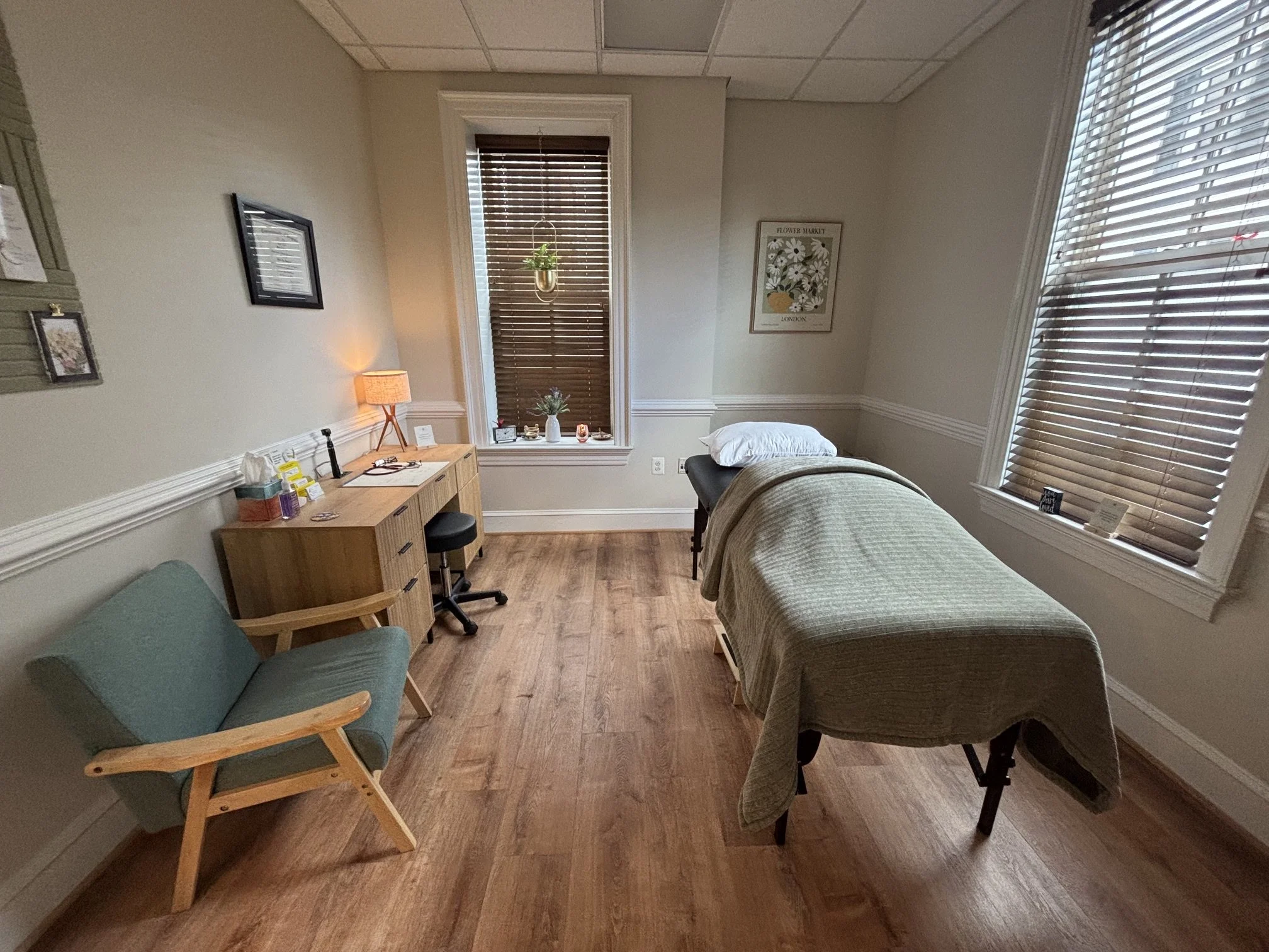 A room with a massage table, a green armchair, a desk with medical supplies, and windows with blinds letting in natural light.