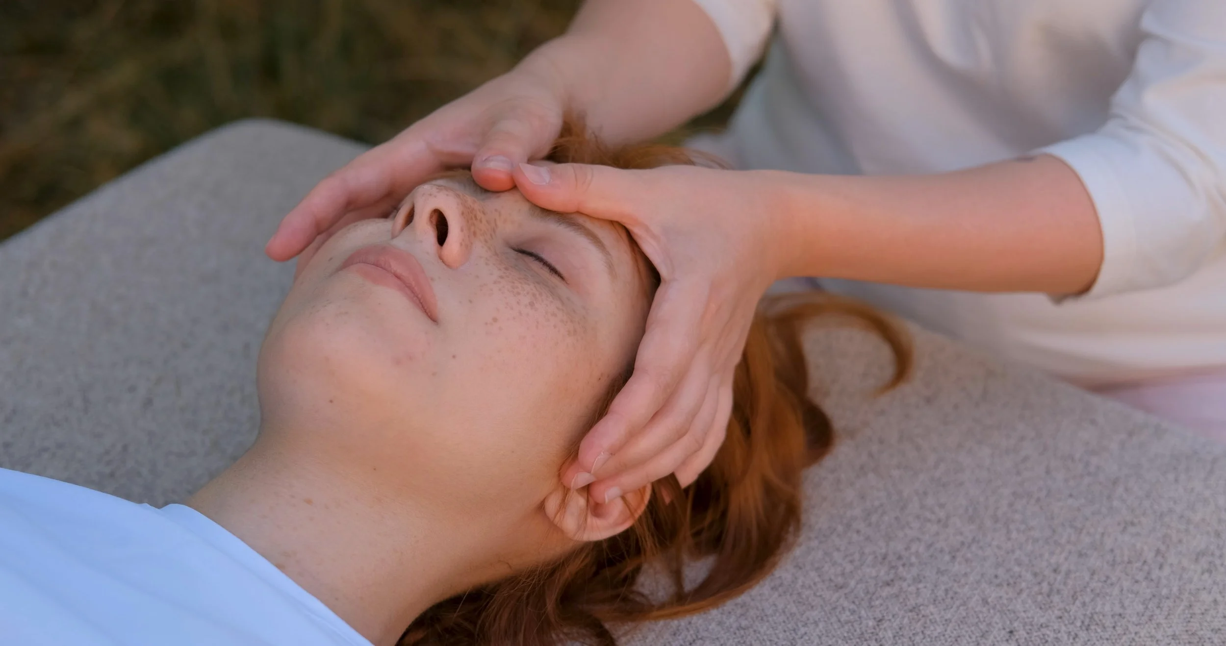 A person with red hair lying on their back receiving a massage or facial treatment, with closed eyes and a relaxed expression, outdoors on a gray surface.