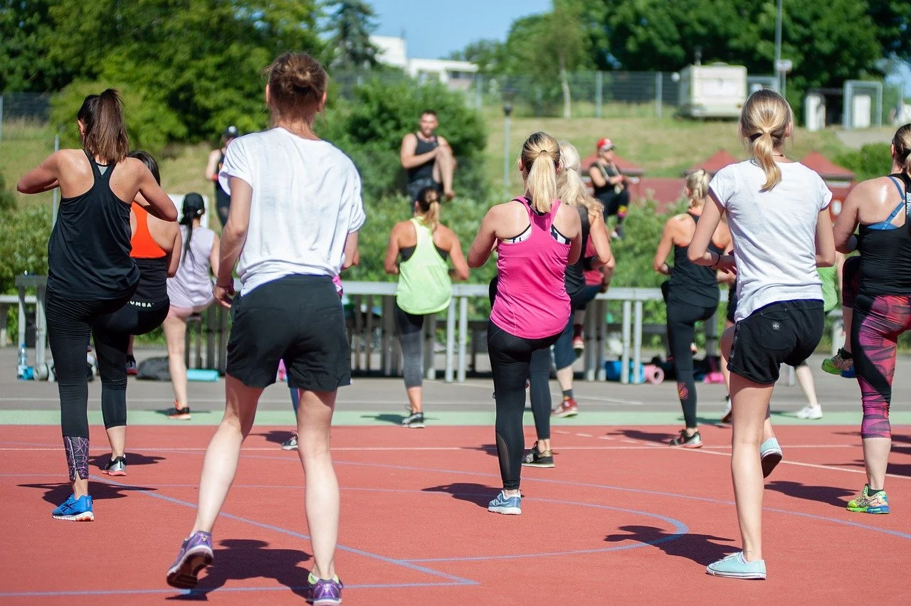 Group of women participating in an outdoor fitness class on a track, doing leg stretches or warm-up exercises.