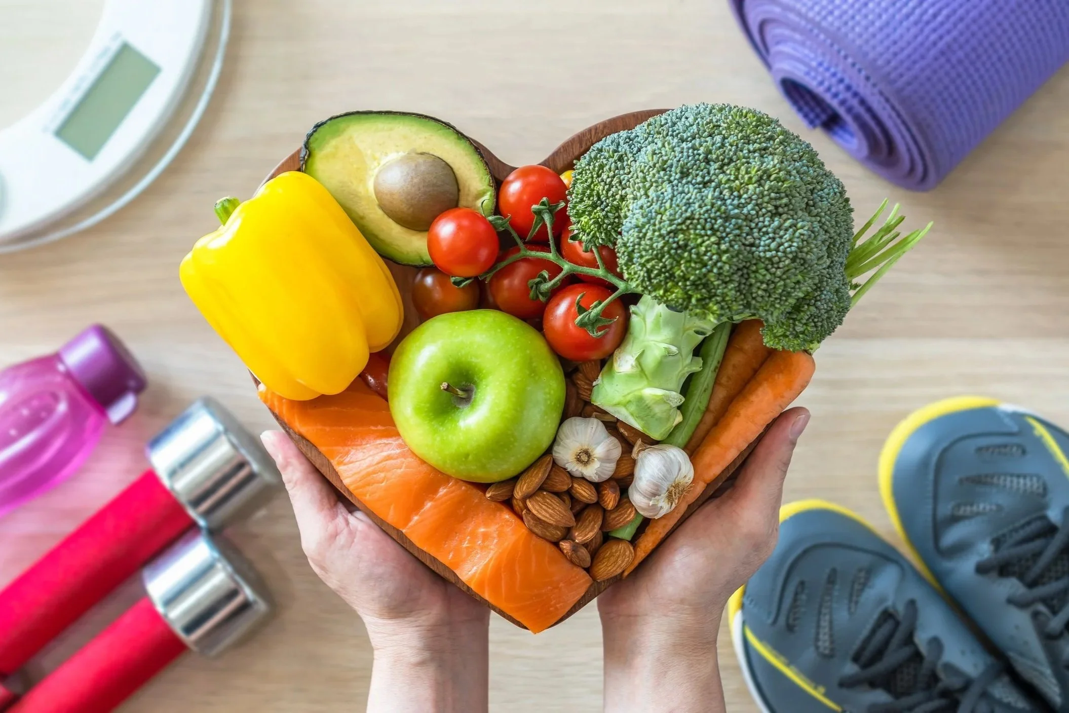 Person holding a heart-shaped wooden bowl filled with fresh vegetables and fruits, including tomatoes, an avocado, a green apple, a yellow bell pepper, broccoli, carrots, garlic, and almonds, with a scale and exercise mat in the background.