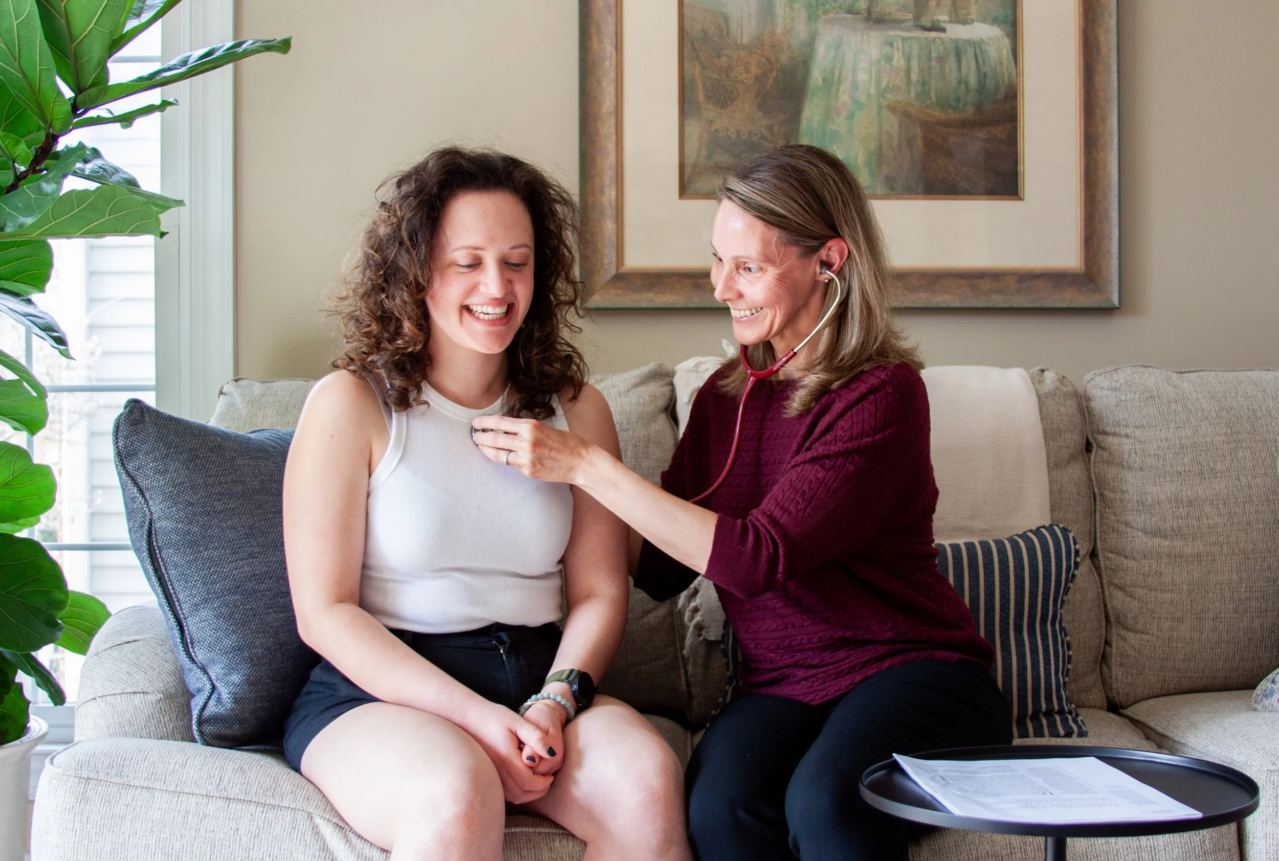 A healthcare professional using a stethoscope to check a young woman's heartbeat during a medical consultation in a living room.