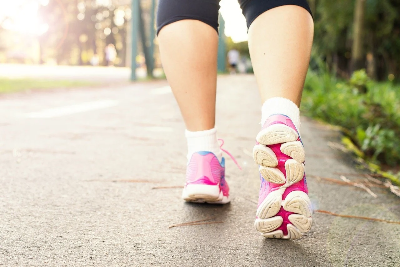 Close-up of a person jogging in a park wearing pink and blue running shoes, white socks, and black shorts on a paved path with greenery beside.