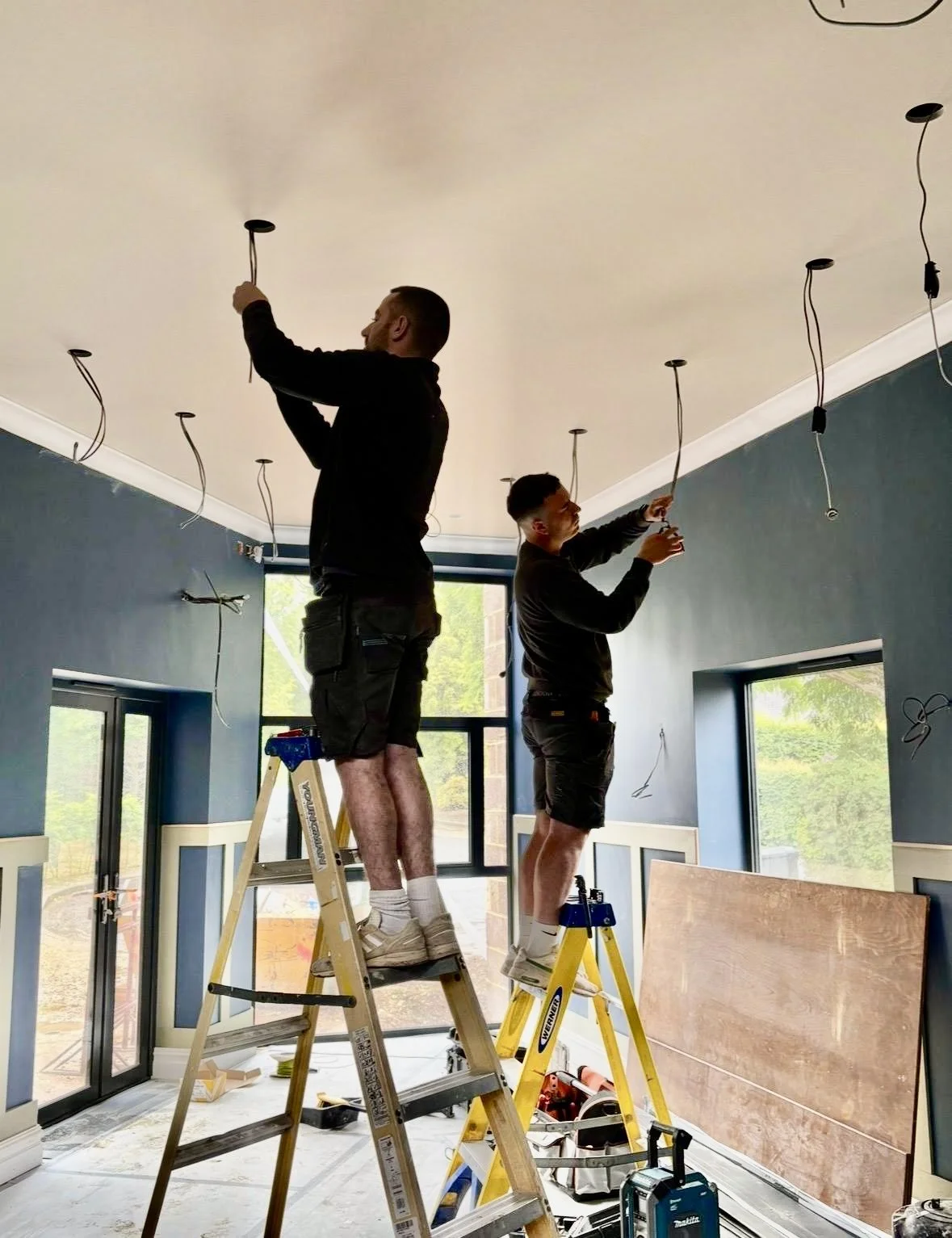 Two men standing on ladders installing recessed lighting fixtures in a ceiling of a room with large windows and blue walls.